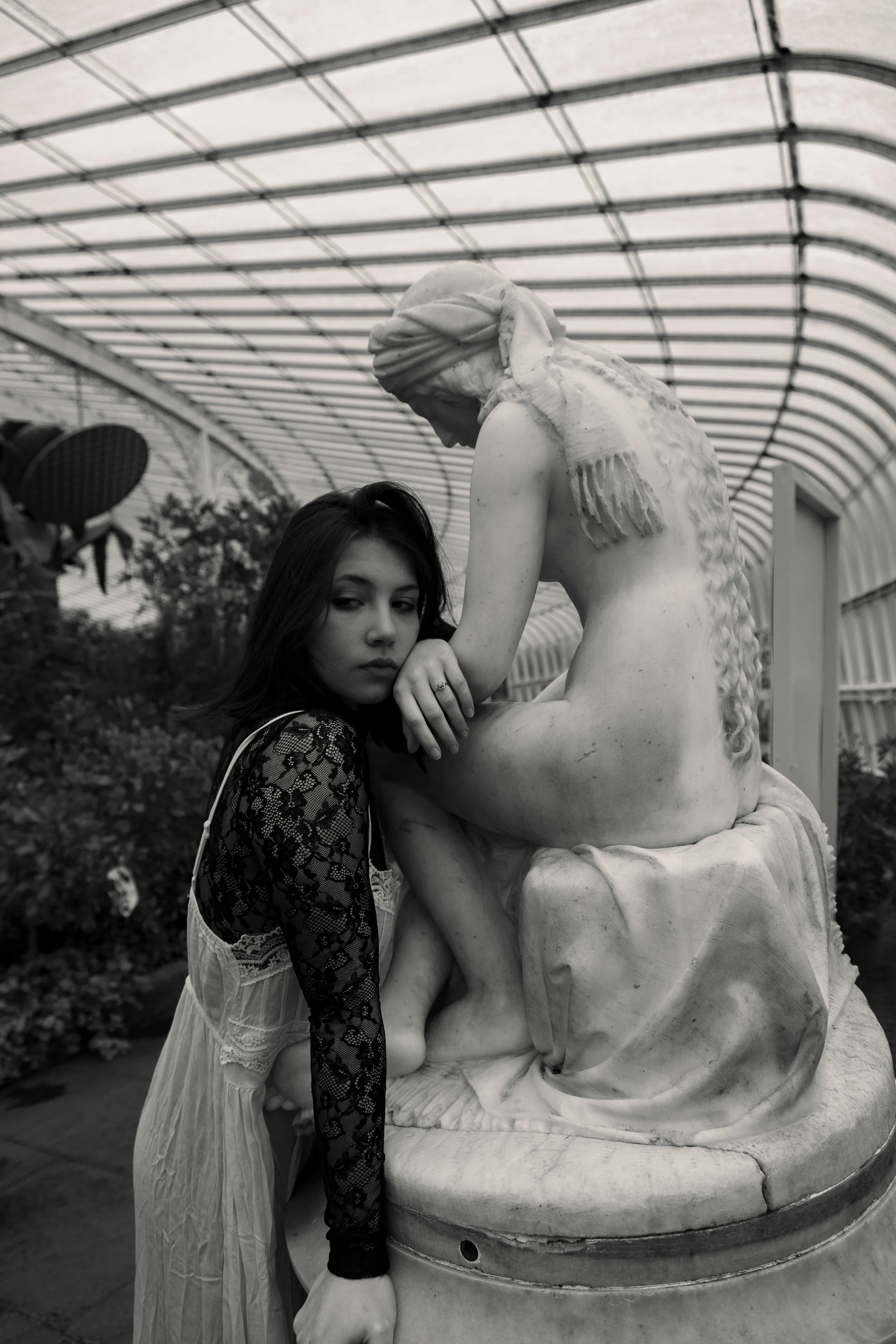 A young woman in a lace dress leaning against a classical marble statue of a seated woman inside a greenhouse or conservatory.