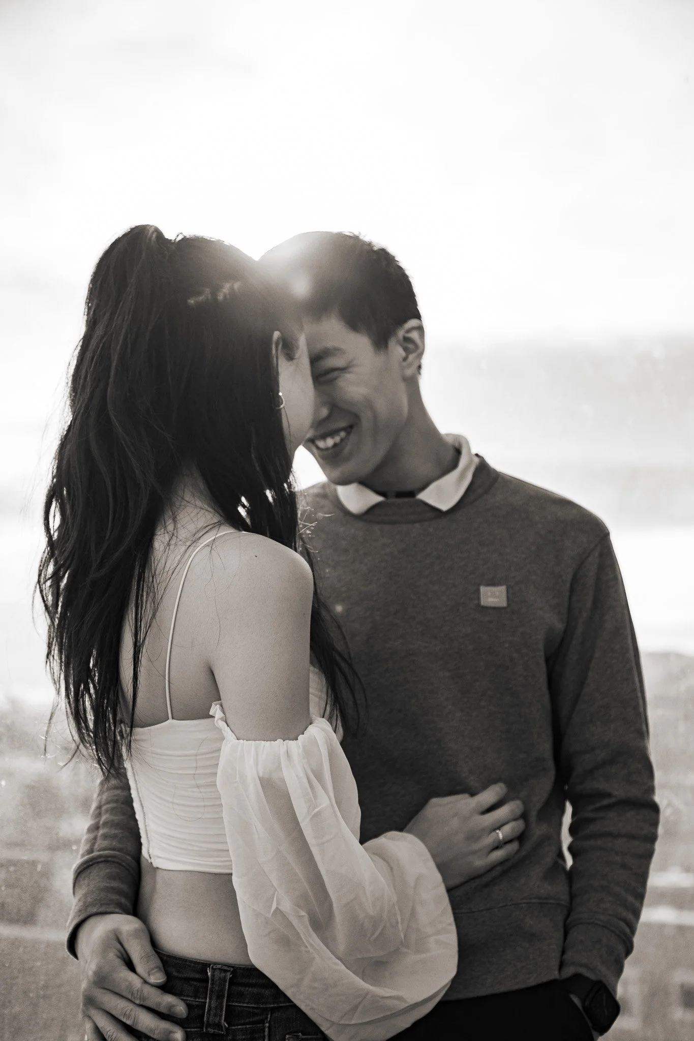 A black and white photo of a smiling young woman and a young man sharing an intimate moment outdoors, their faces close together, showing affection.