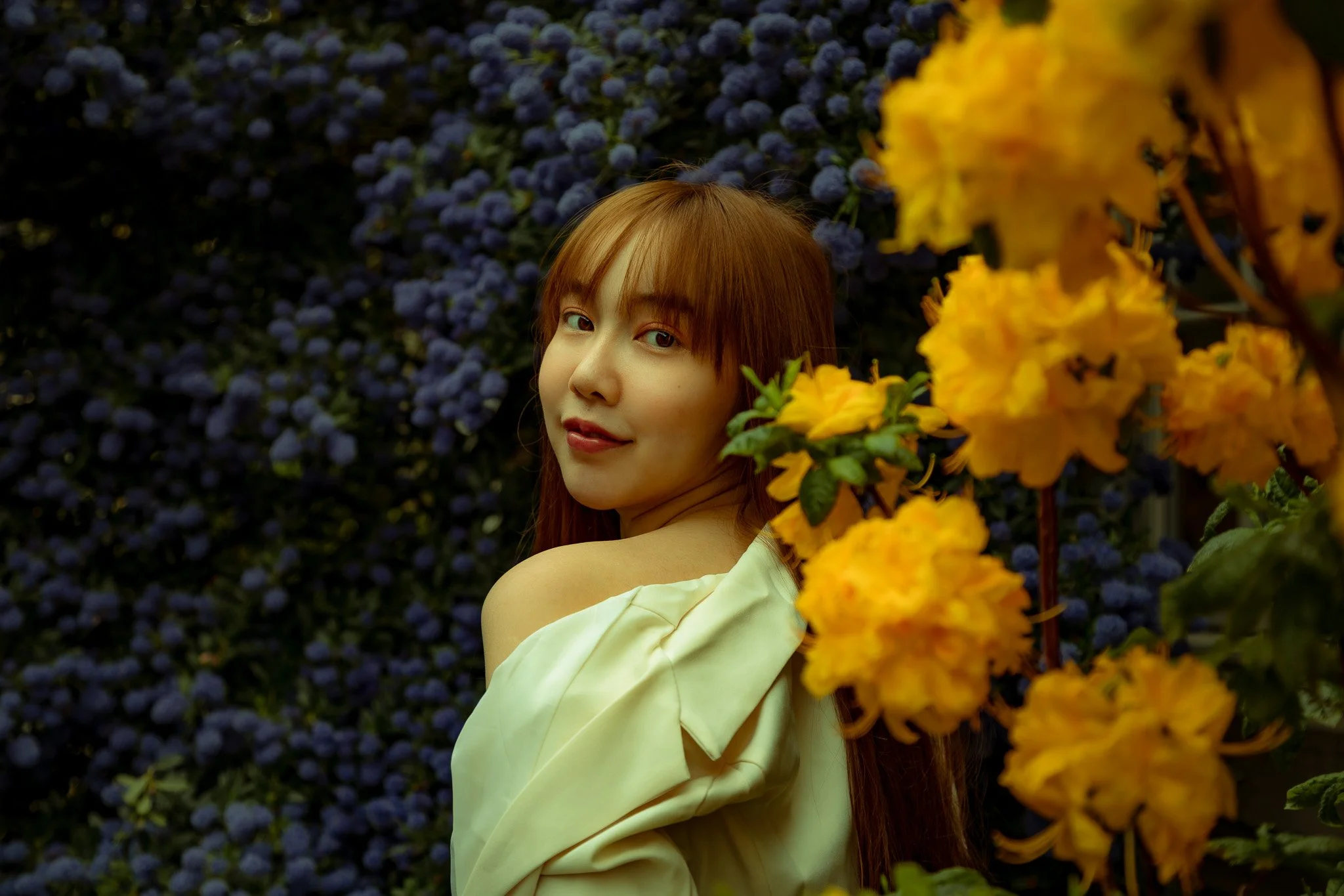 A woman with long, brown hair looking over her shoulder in front of a backdrop of purple and yellow flowers.