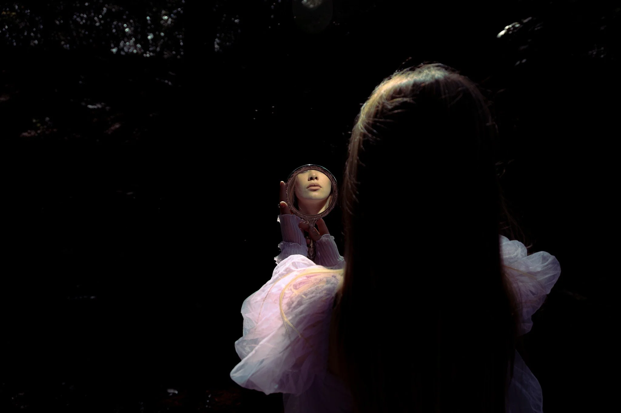 A woman with long hair, dressed in a white outfit, holds a small mirror reflecting her face against a dark background.