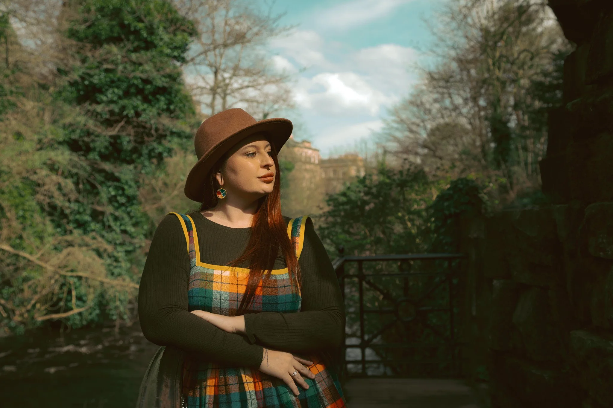 A woman in a plaid dress and brown hat standing outdoors near a river and trees.