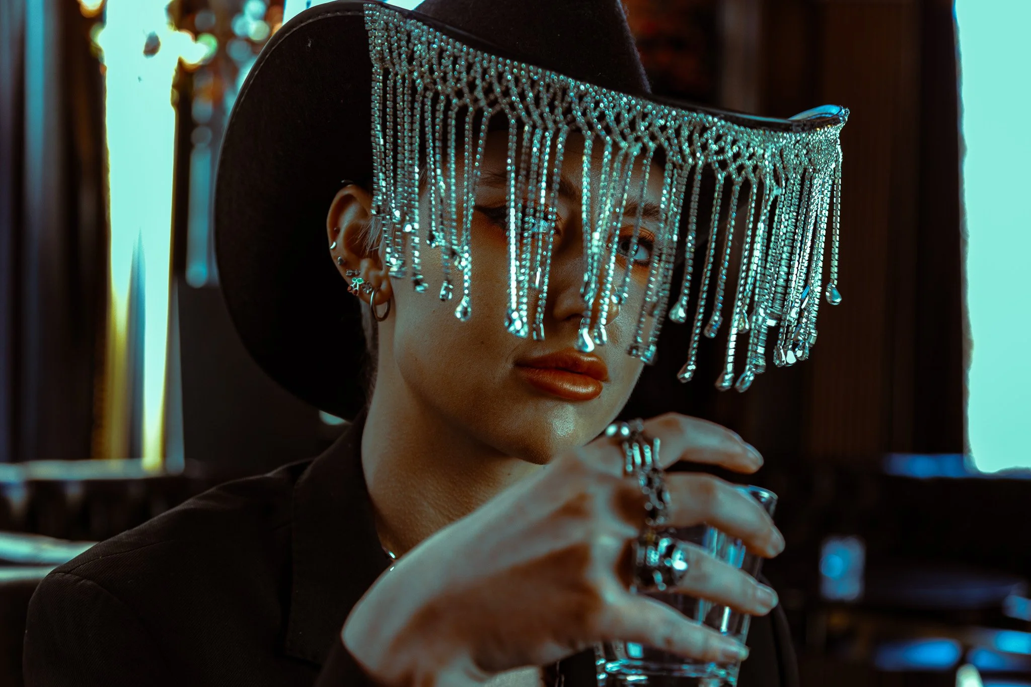 A woman wearing a large black hat with a shiny, bejeweled fringe, holding a glass of water, with dark background and warm lighting.