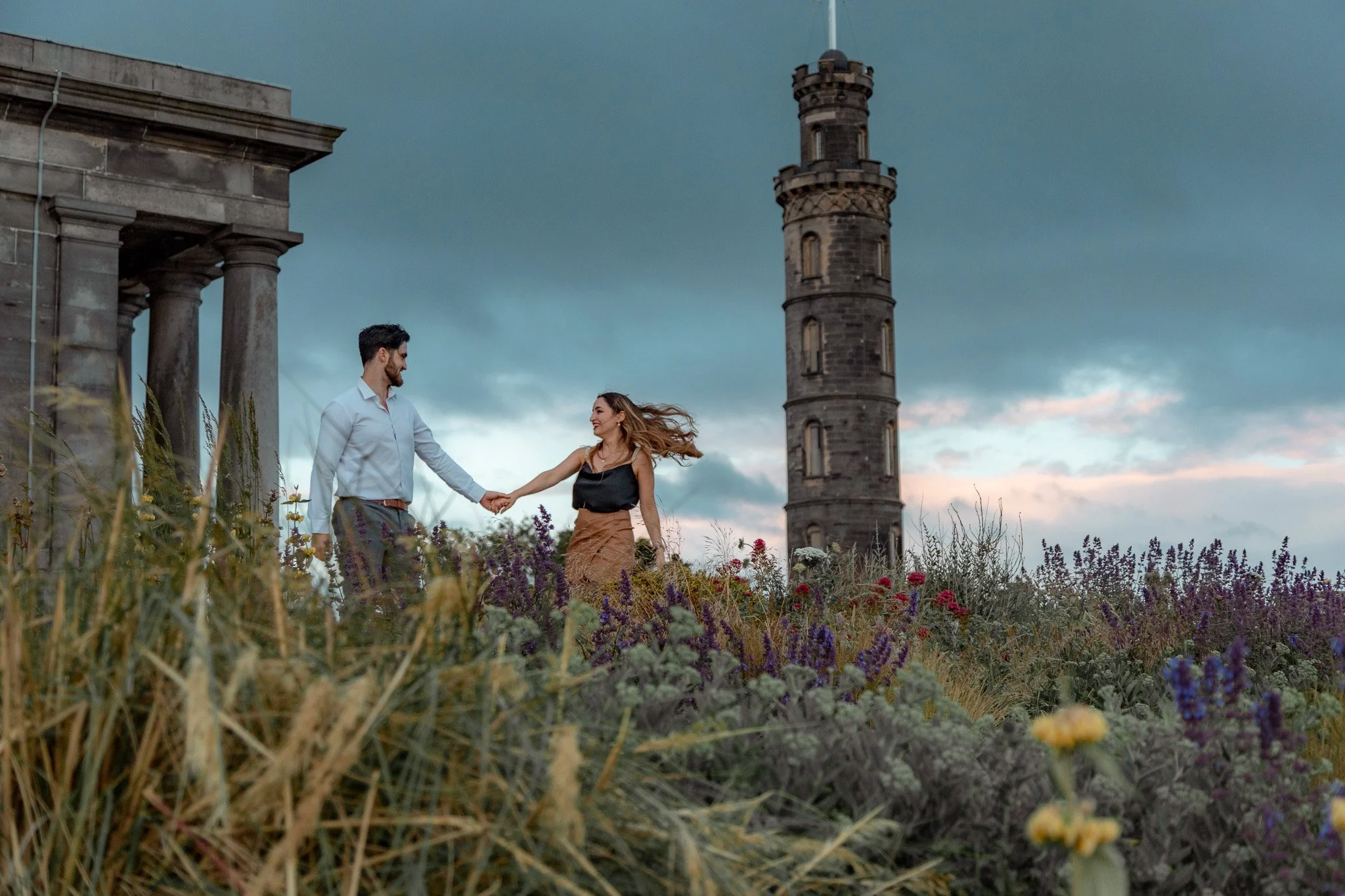 A man and woman holding hands and smiling in a field of wildflowers with a tall, round stone tower and a cloudy sky in the background.