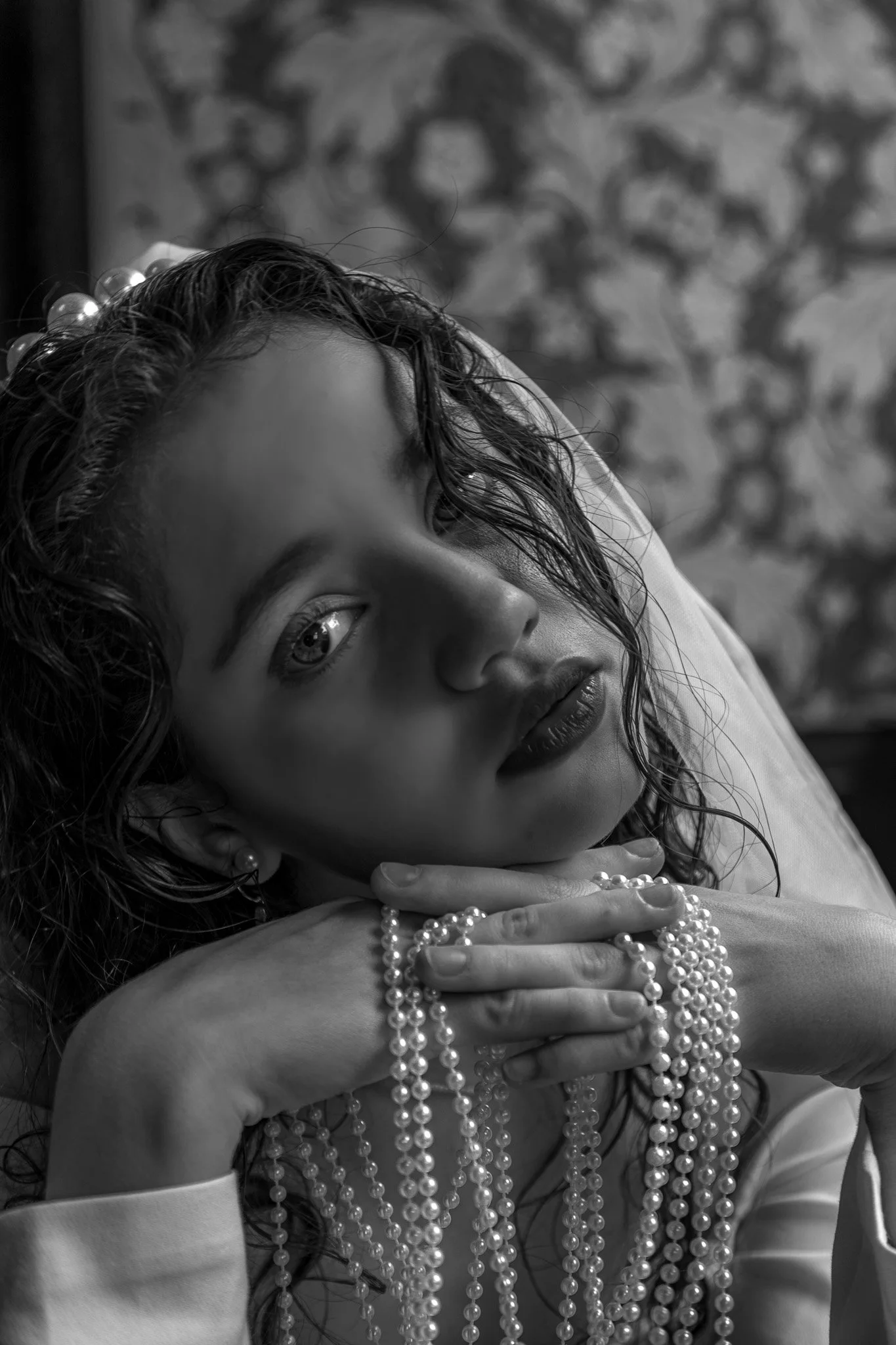 Black and white portrait of a woman with curly hair, holding multiple pearl necklaces, resting her chin on her hands, with a patterned background.