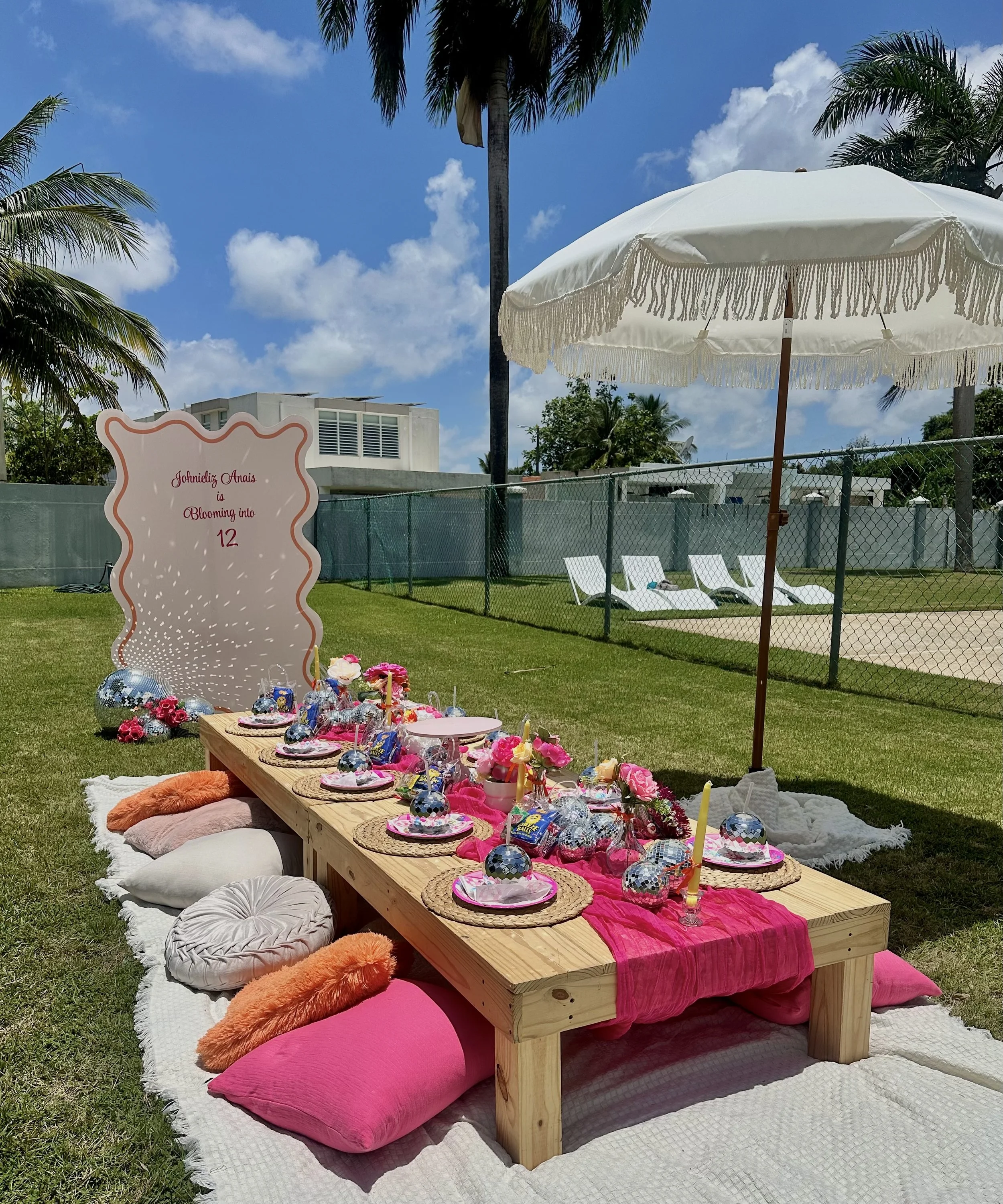 Decoración para fiesta de cumpleaños al aire libre con mesa baja de madera, cojines y mantas rosas y naranjas, sobre césped junto a una cerca, con sombrilla blanca y en el fondo palmeras y cielo azul.