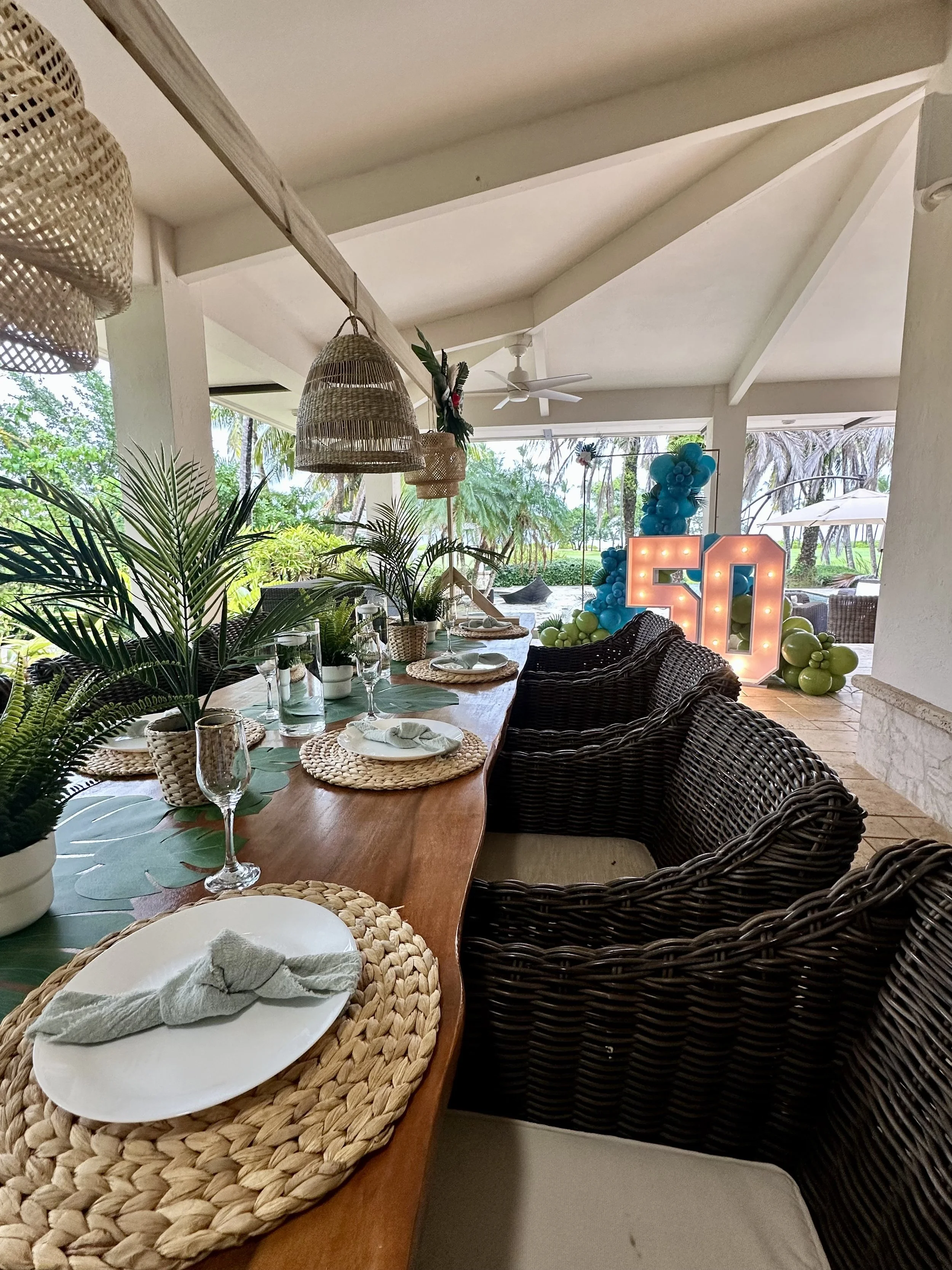 Mesa decorada para celebración del 50 cumpleaños con platos, copas y plantas verdes, en un espacio abierto con vista a plantas y decoración con luces y globos de colores.