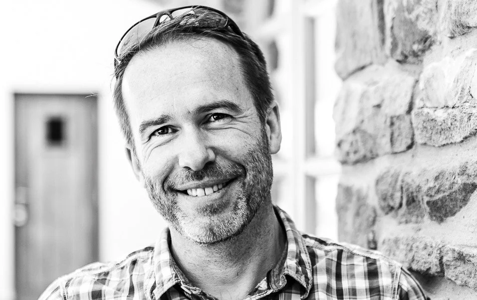 A photograph of a man smiling at the camera. He wears sunglasses on his head and has a short beard