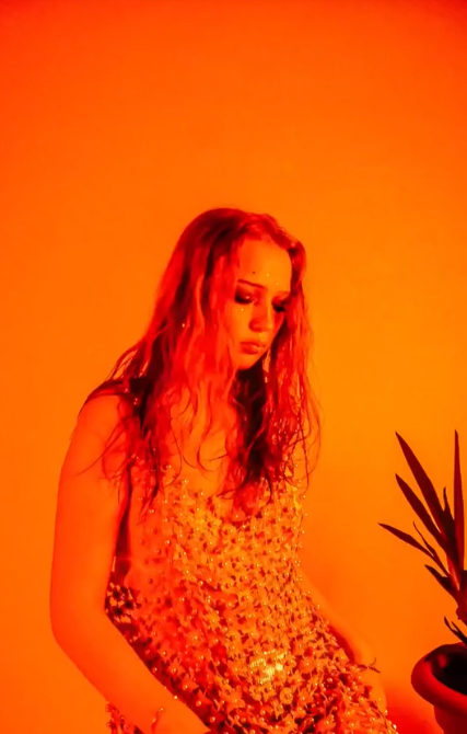 A woman with long, curly red hair wearing a sparkling, gold sequin dress, sitting next to a plant against an orange background.