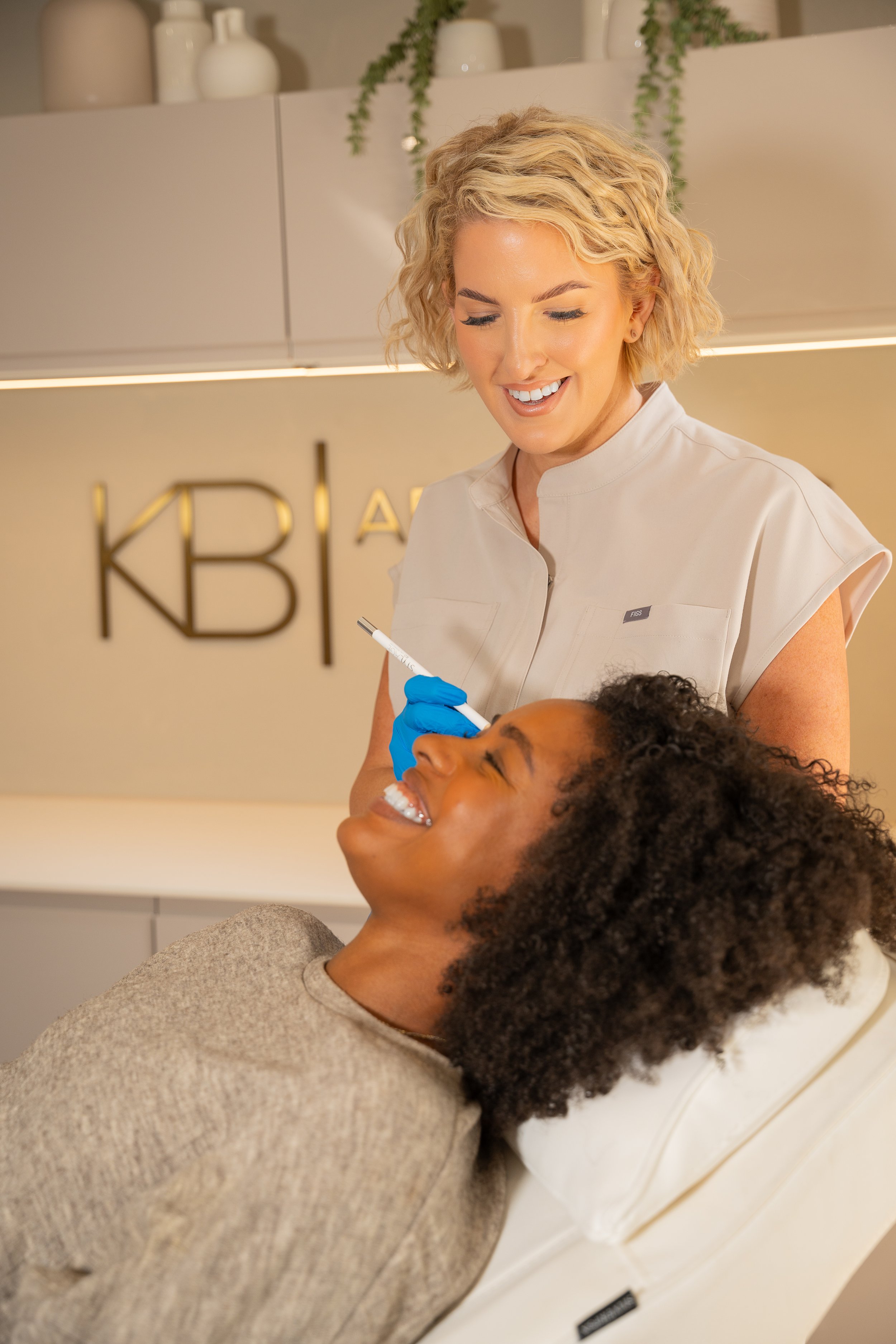 A woman lying on a medical examination chair smiling while a healthcare professional, possibly a nurse or doctor, prepares to give her a facial or cosmetic treatment with a syringe. The healthcare worker wears blue gloves and the environment looks like a modern clinic.