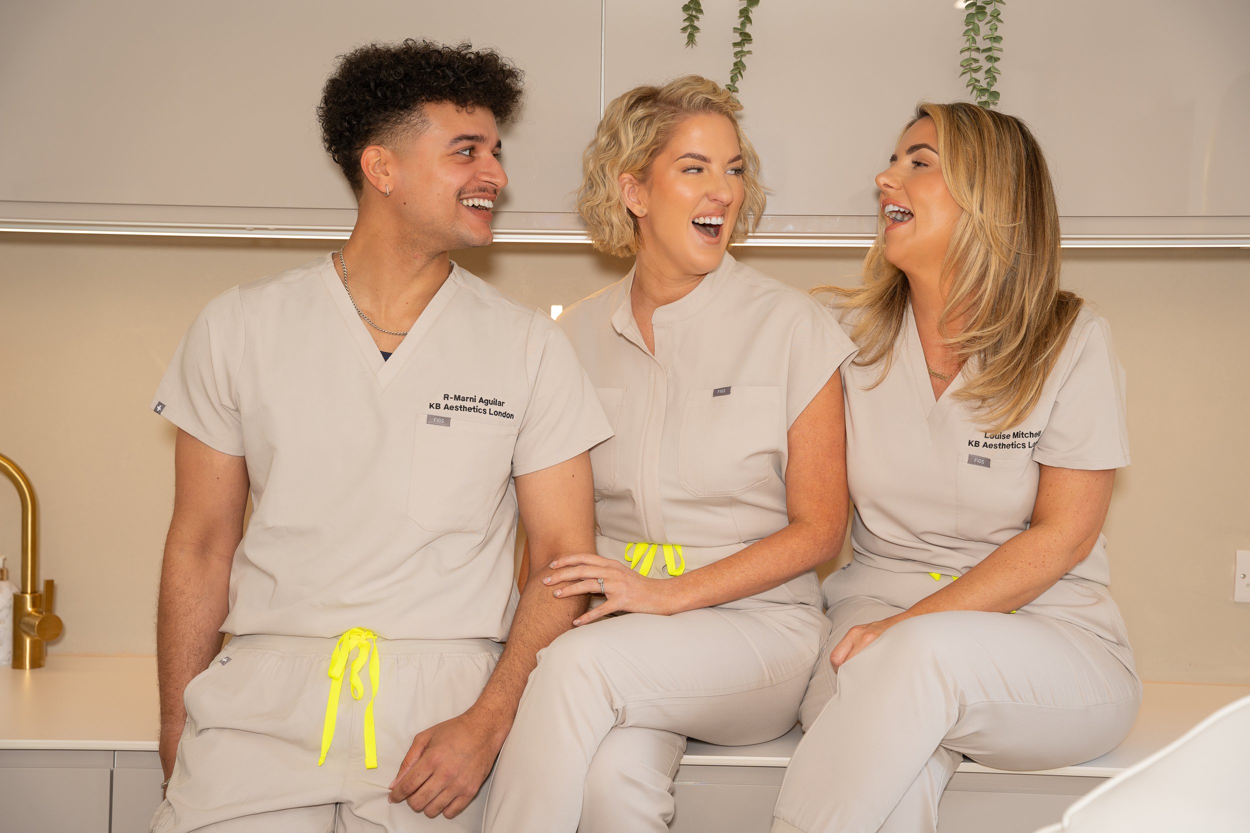 Three healthcare professionals in matching beige scrubs sitting on a counter, smiling and engaging in conversation.