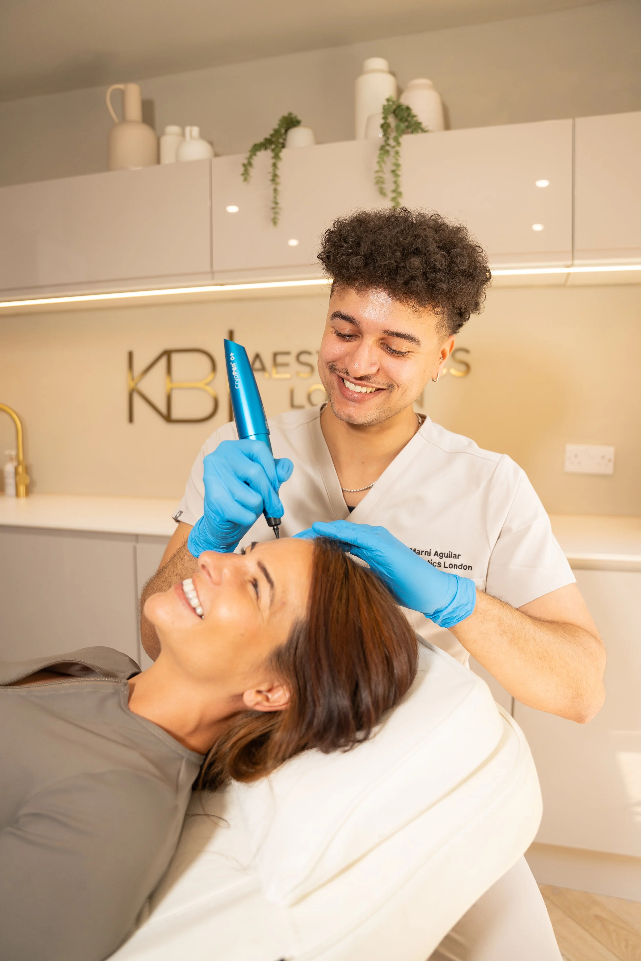 A smiling woman lying on a treatment bed getting a cosmetic procedure on her forehead from a male aesthetician wearing gloves in a modern clinic.