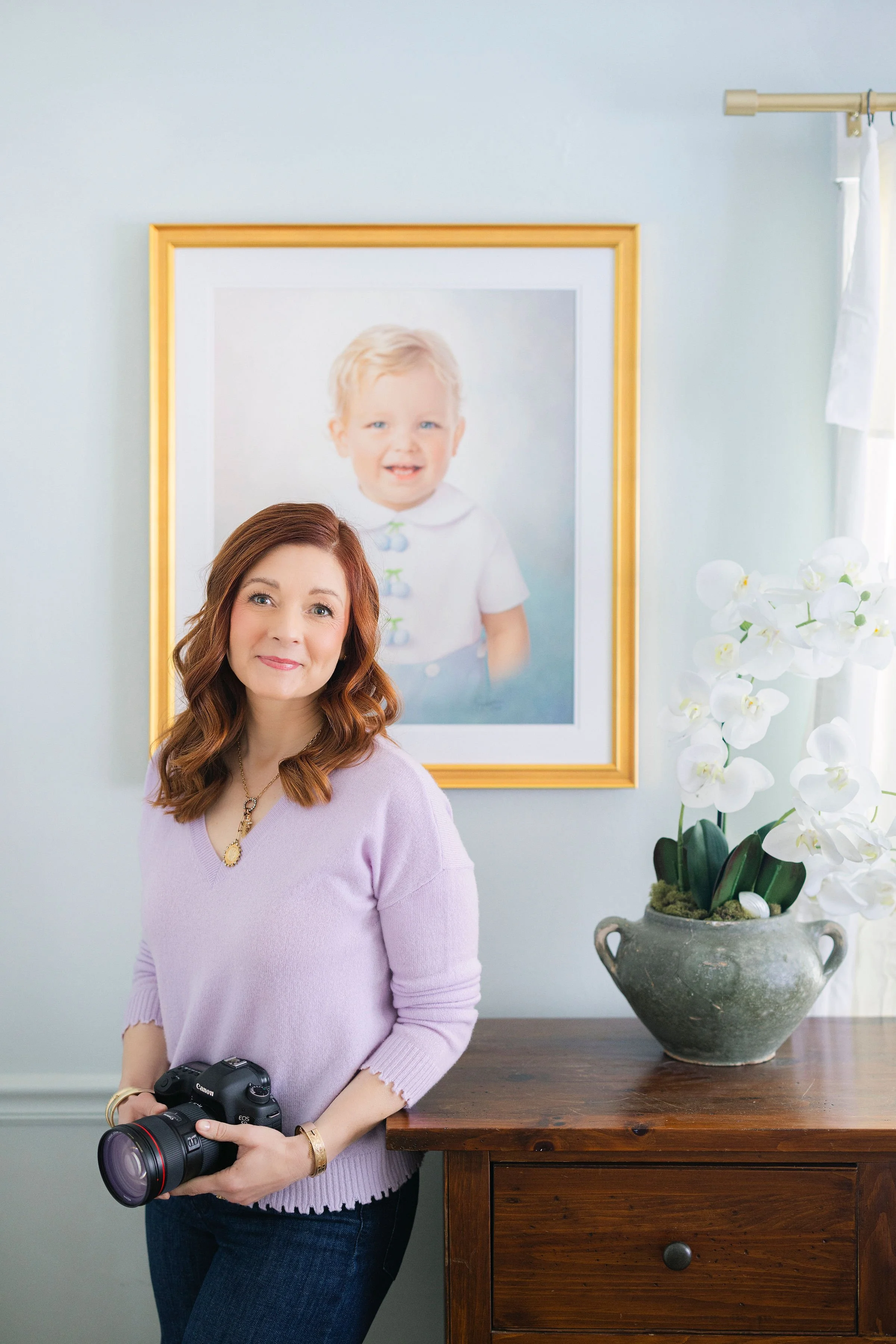 Shana Watkins holding her camera beside a framed children’s heirloom portrait displayed in a home.