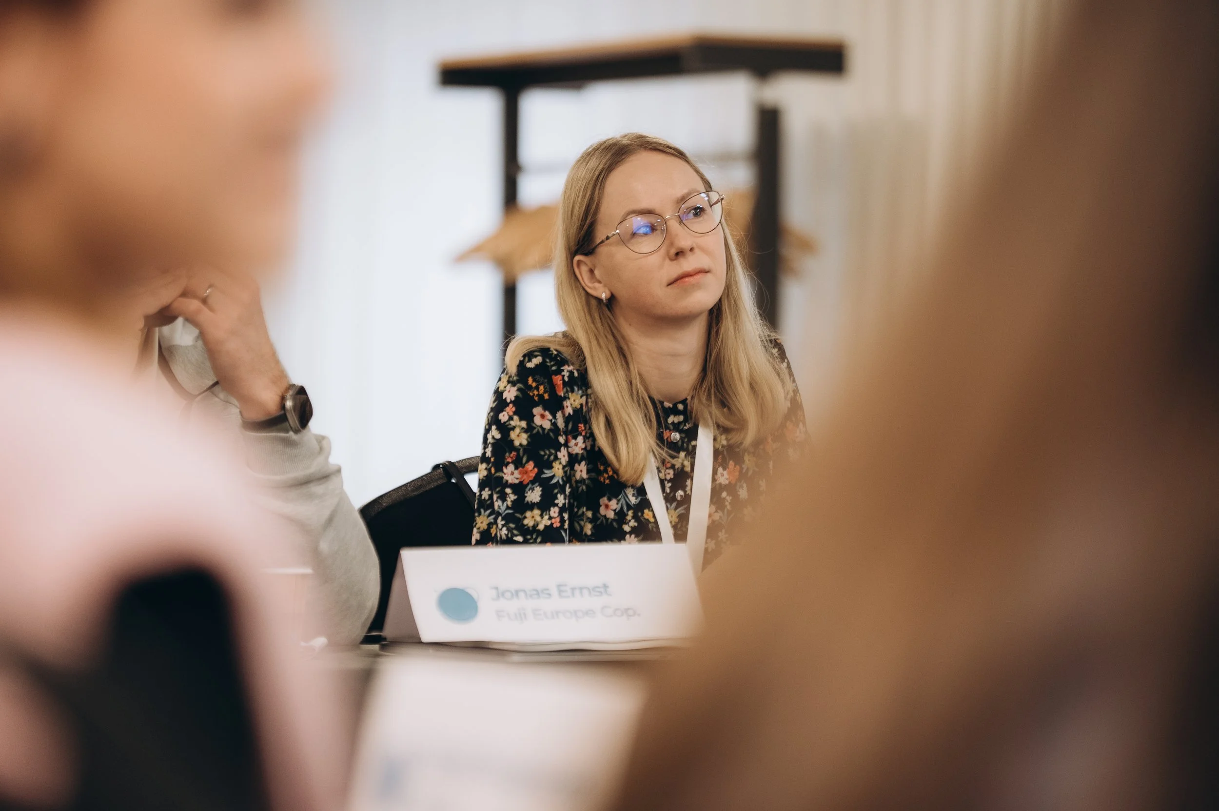 A woman with blonde hair and glasses sitting at a table during a meeting, with a nameplate that reads 'Jonas Ernst, Fuji Europe Cop.' in front of her.