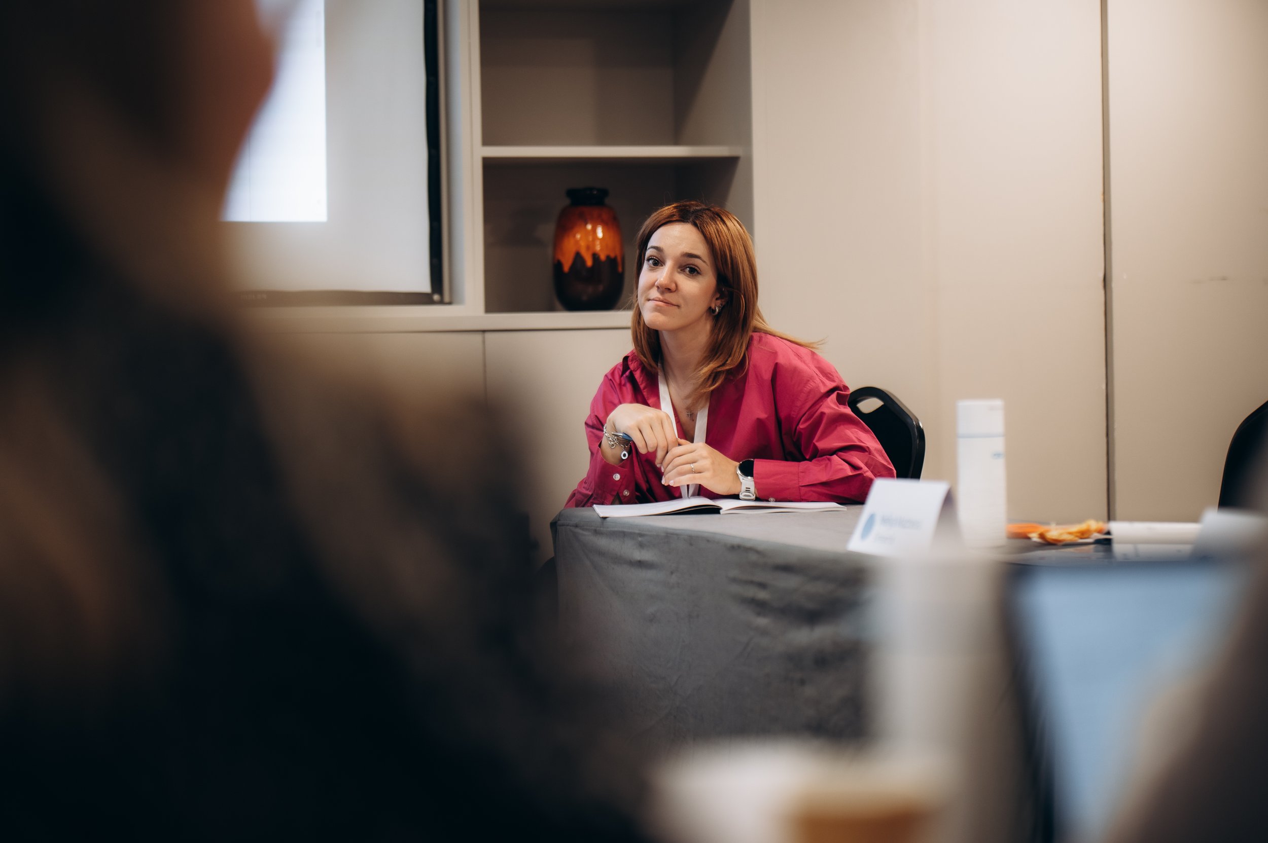 A young woman with red hair in a pink shirt seated at a conference table, looking attentively during a meeting, with a notebook and some items on the table.