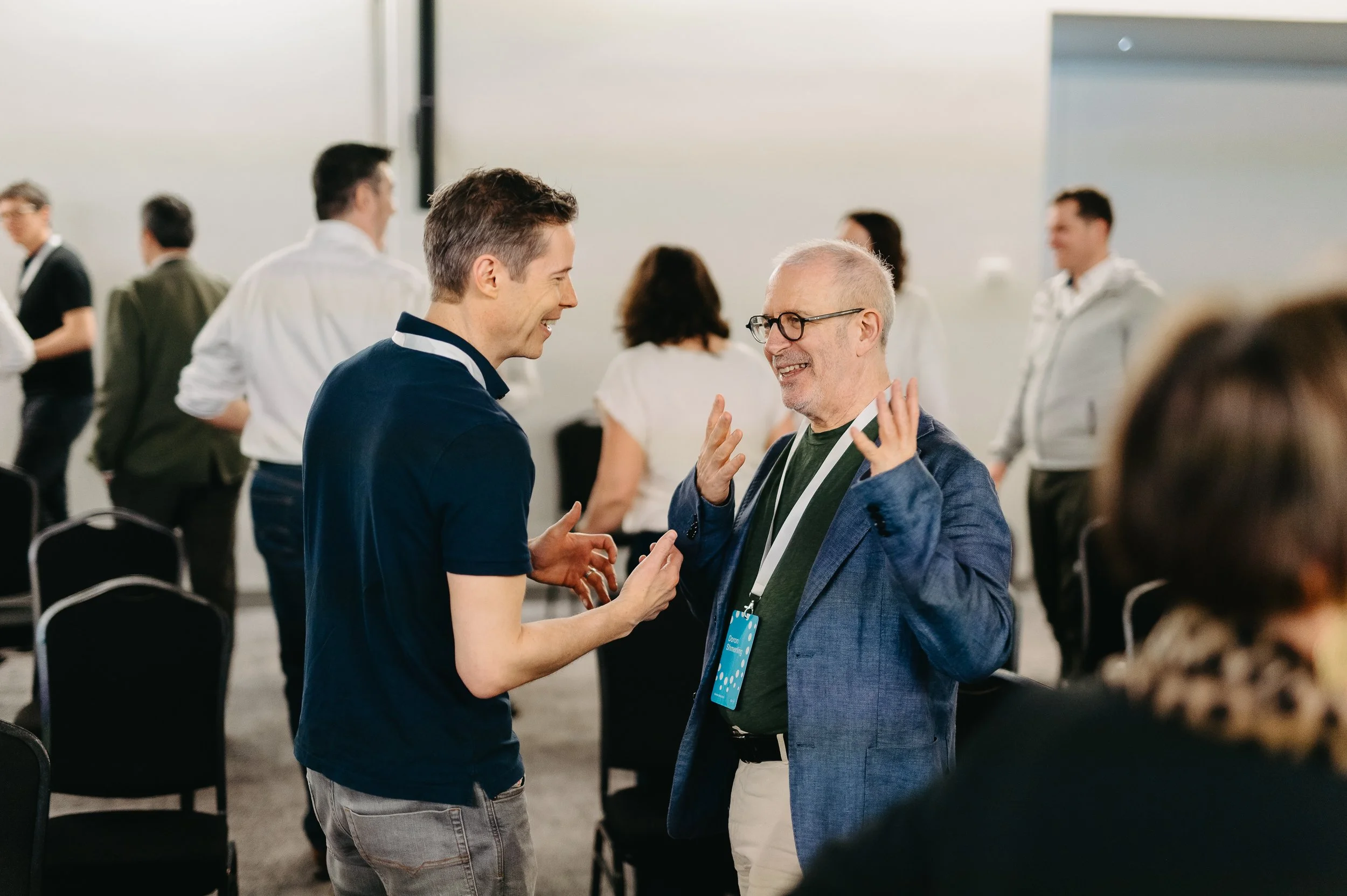 Two men engaging in a lively conversation and smiling in a conference or networking event with other attendees in the background.