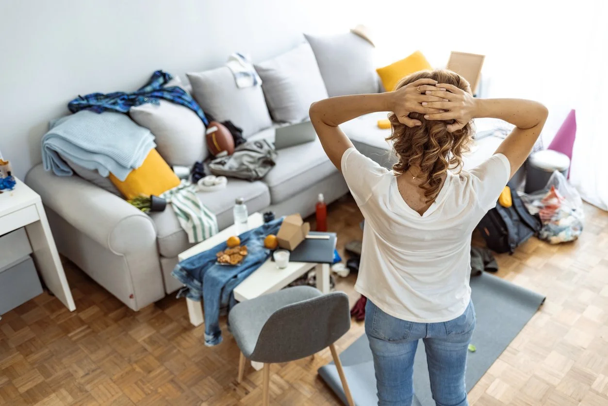 erson stands in a messy living room with hands on head, feeling overwhelmed by undone chores—illustrating how missed acts of service create stress in relationships.