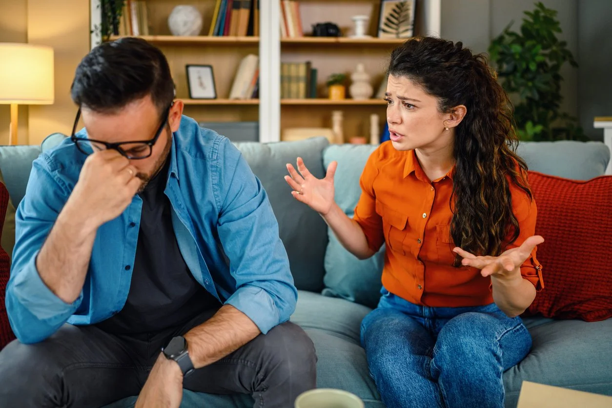 Couple on a sofa during an argument—woman expressing frustration, man withdrawn and stressed—illustrating unresolved conflict and unfinished business.