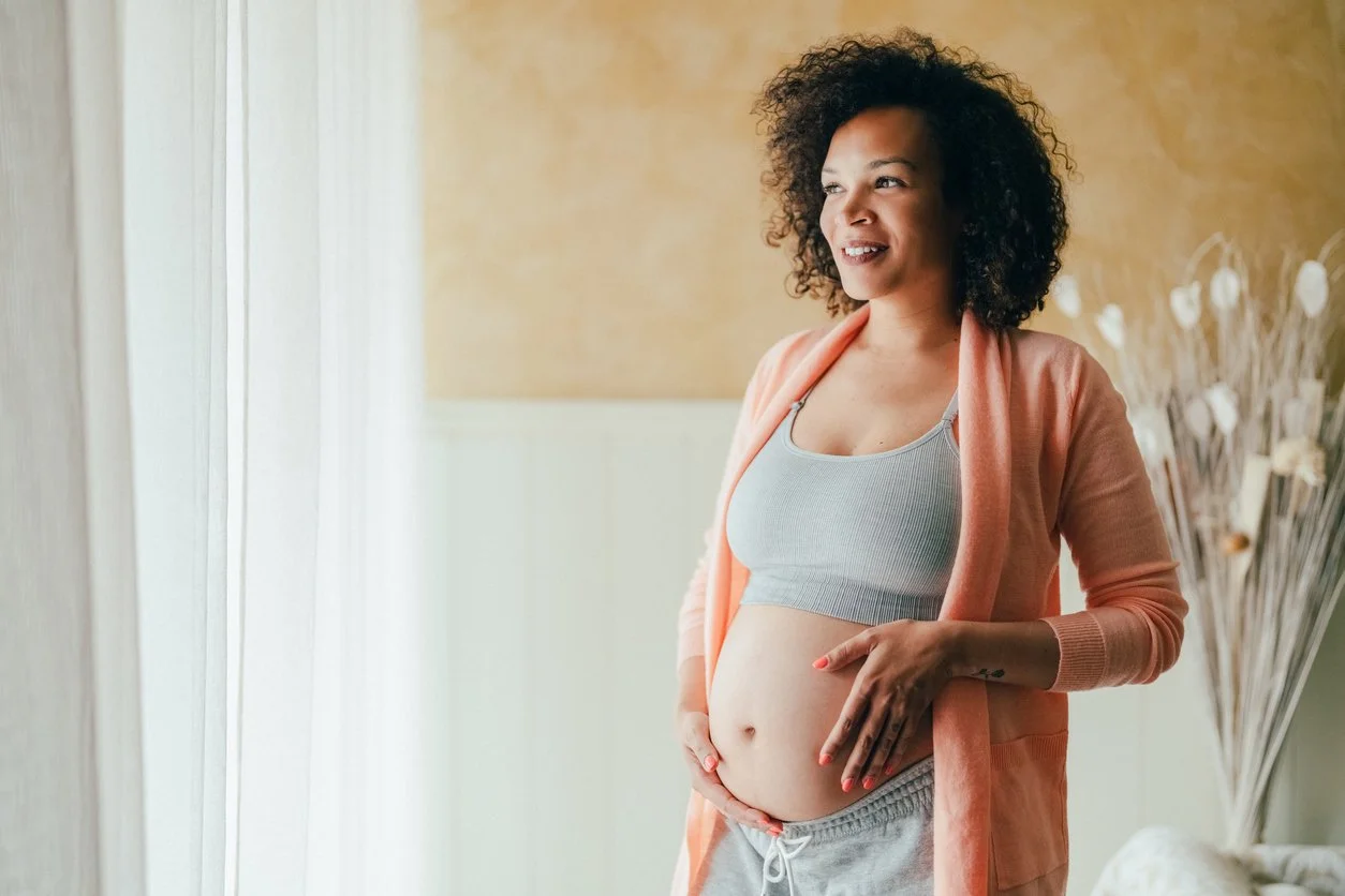 Pregnant woman standing by a window with hands on her belly, looking calm and reflective in the final trimester.