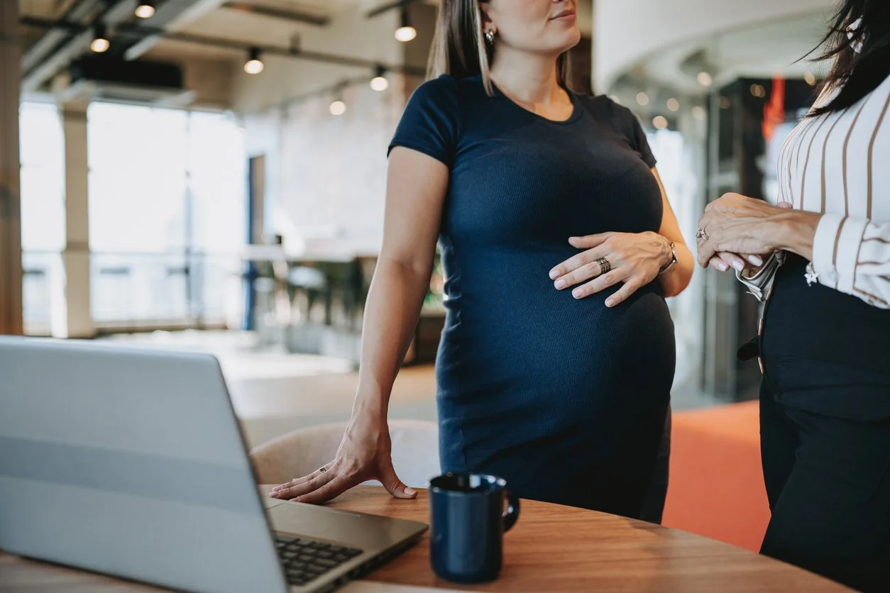 Pregnant person standing beside a desk while talking with a colleague, reflecting work adjustments and boundaries in the third trimester.