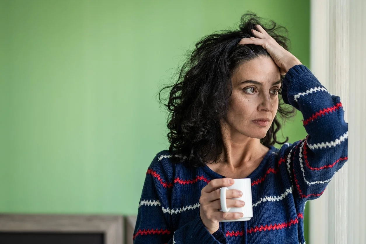 Woman standing by a window, hand in hair and holding a mug, looking worried—symbolizing unresolved feelings in a relationship.