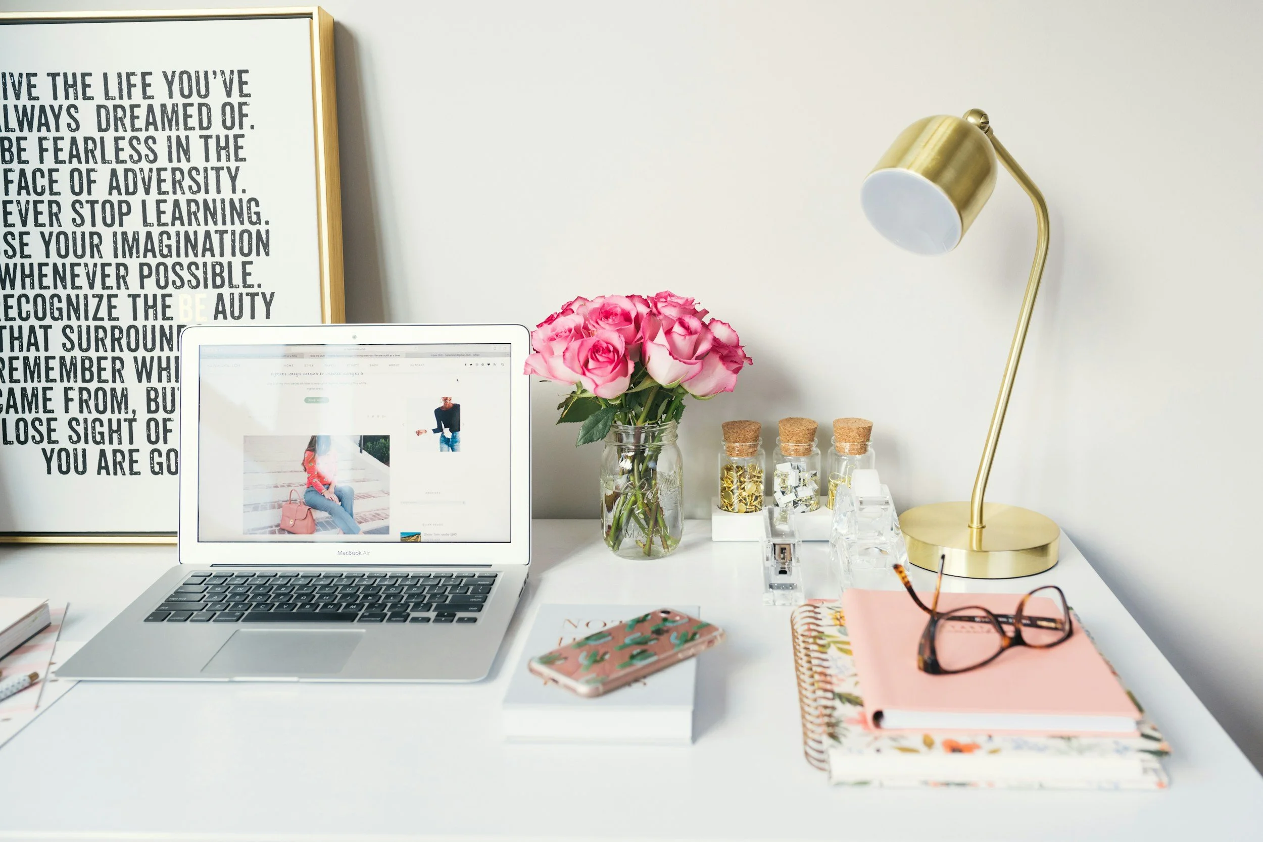 White desk with a MacBook, pink roses in a glass jar, notebooks, glasses, and a gold desk lamp. A framed motivational poster hangs on the wall behind the desk.