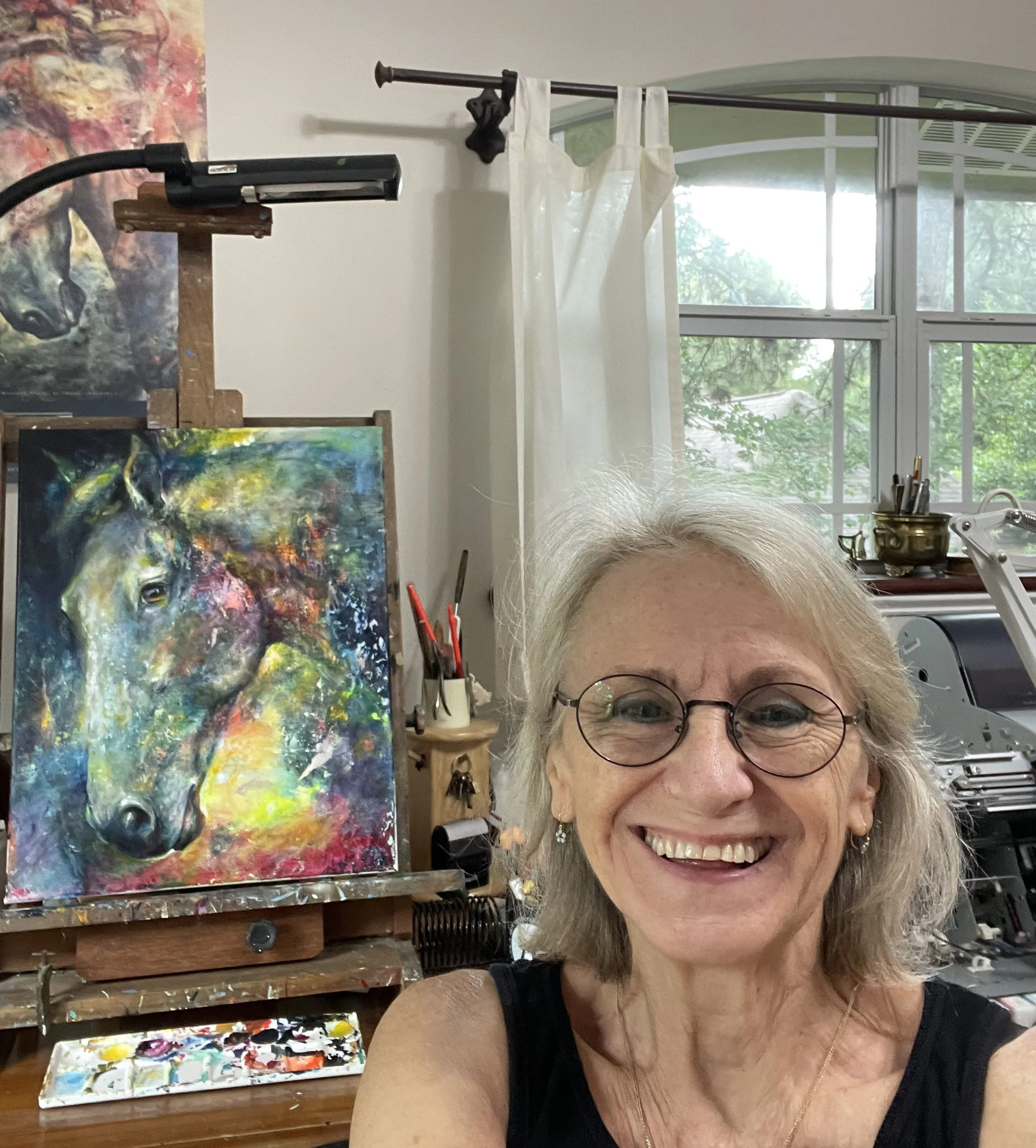 A smiling woman with glasses and earrings in an art studio, with a colorful horse portrait painting on an easel behind her.