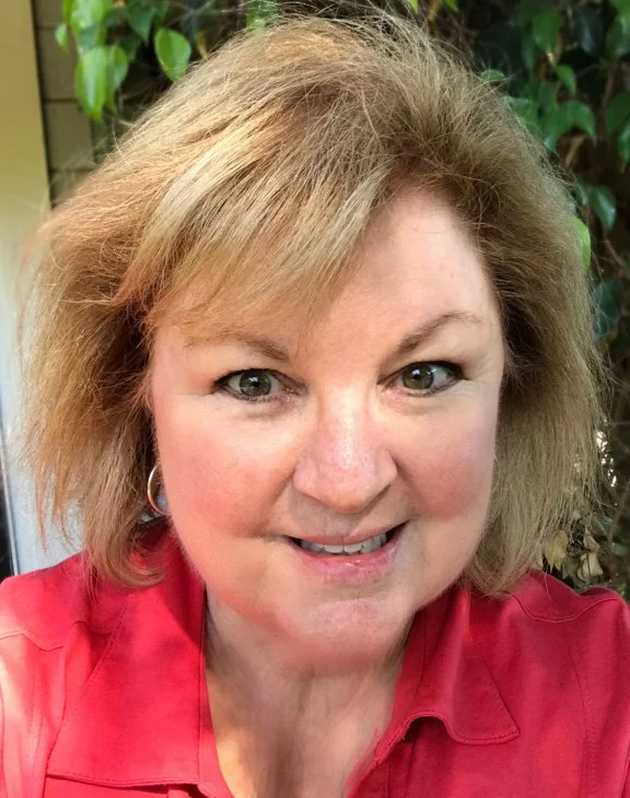 A woman with blonde hair and blue eyes smiling at the camera, wearing a red shirt, with green foliage in the background.