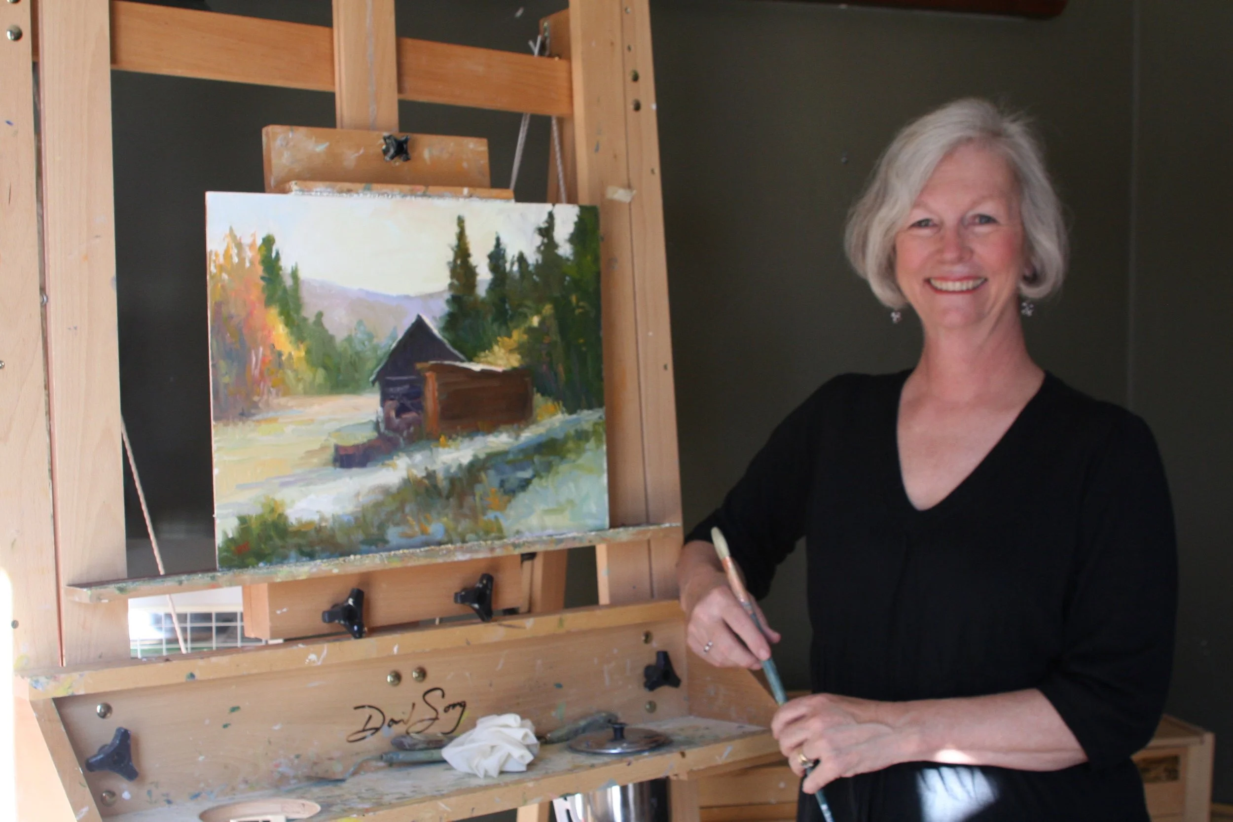Amy Evans standing next to her easel in the studio with a landscape painting on the easel filled with sunlight and beauty