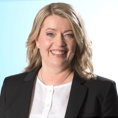 A woman with blonde hair, smiling, wearing a black blazer and white shirt, against a light blue background.