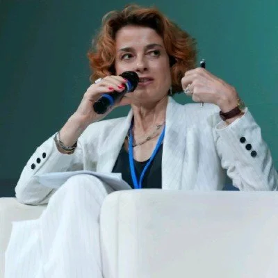 Woman with short curly red hair speaking into a microphone while sitting on a white couch, holding papers in her lap.