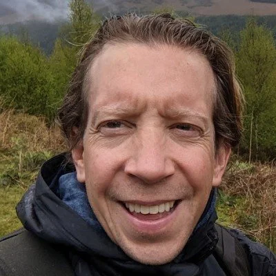 Close-up of a smiling man outdoors with green trees and hills in the background.