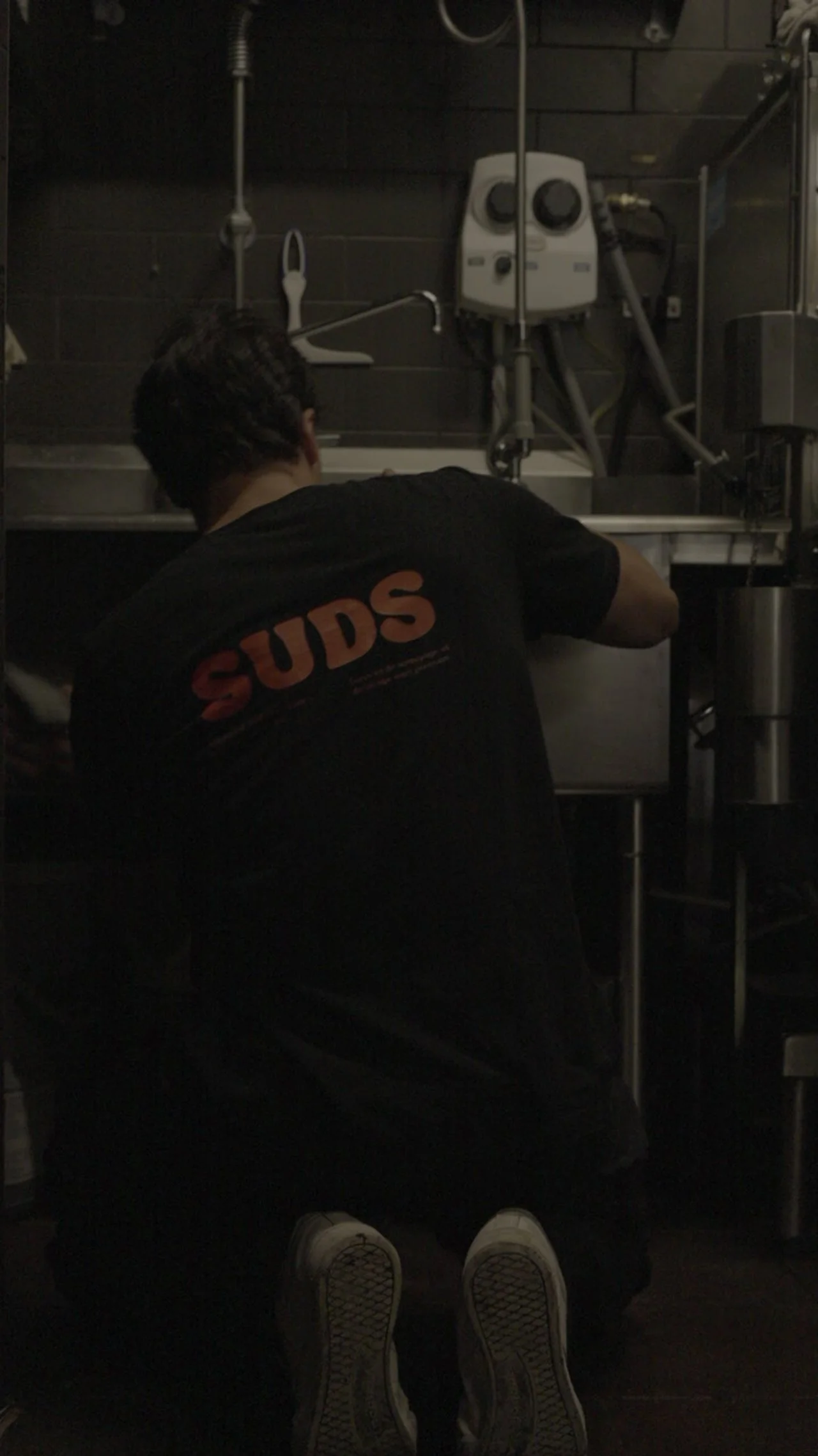 A person wearing a black shirt with 'SUDS' on the back, kneeling in a dimly lit kitchen or bar area, working with stainless steel equipment.