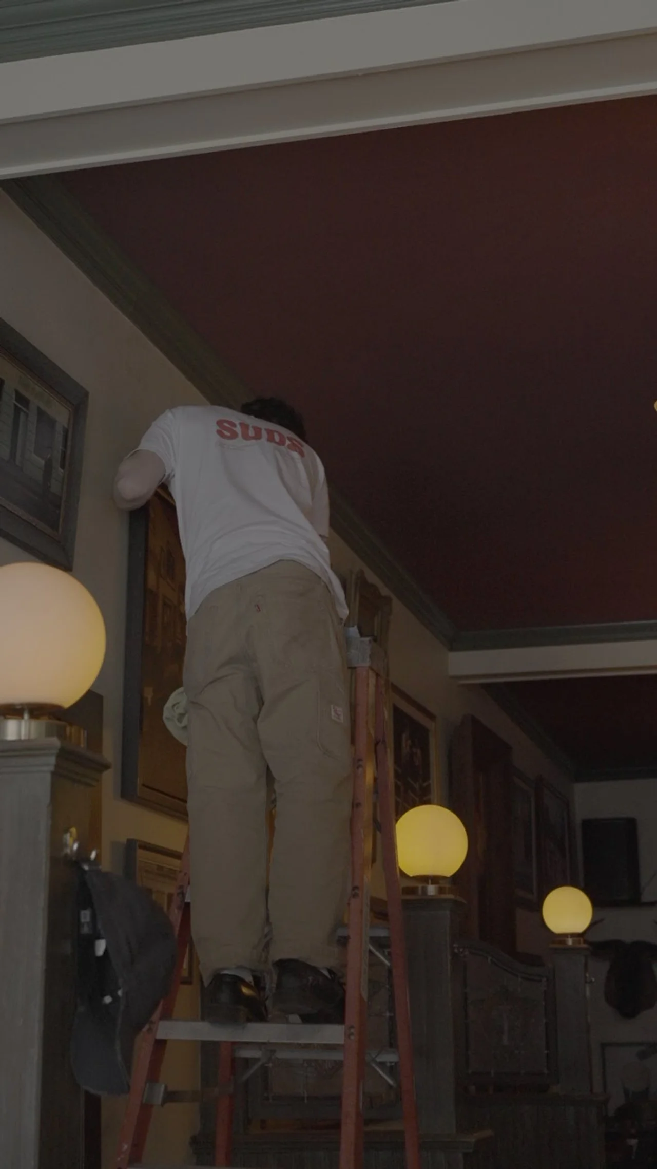 A person standing on a ladder reaching up to a ceiling, wearing a white T-shirt and khaki pants in a dimly lit indoor space with wall art and lamps.