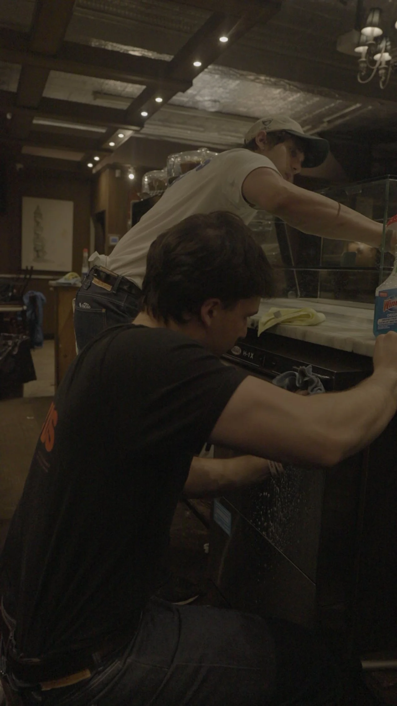 Two men working behind a bar counter in a restaurant or bar, with one cleaning and the other preparing drinks.