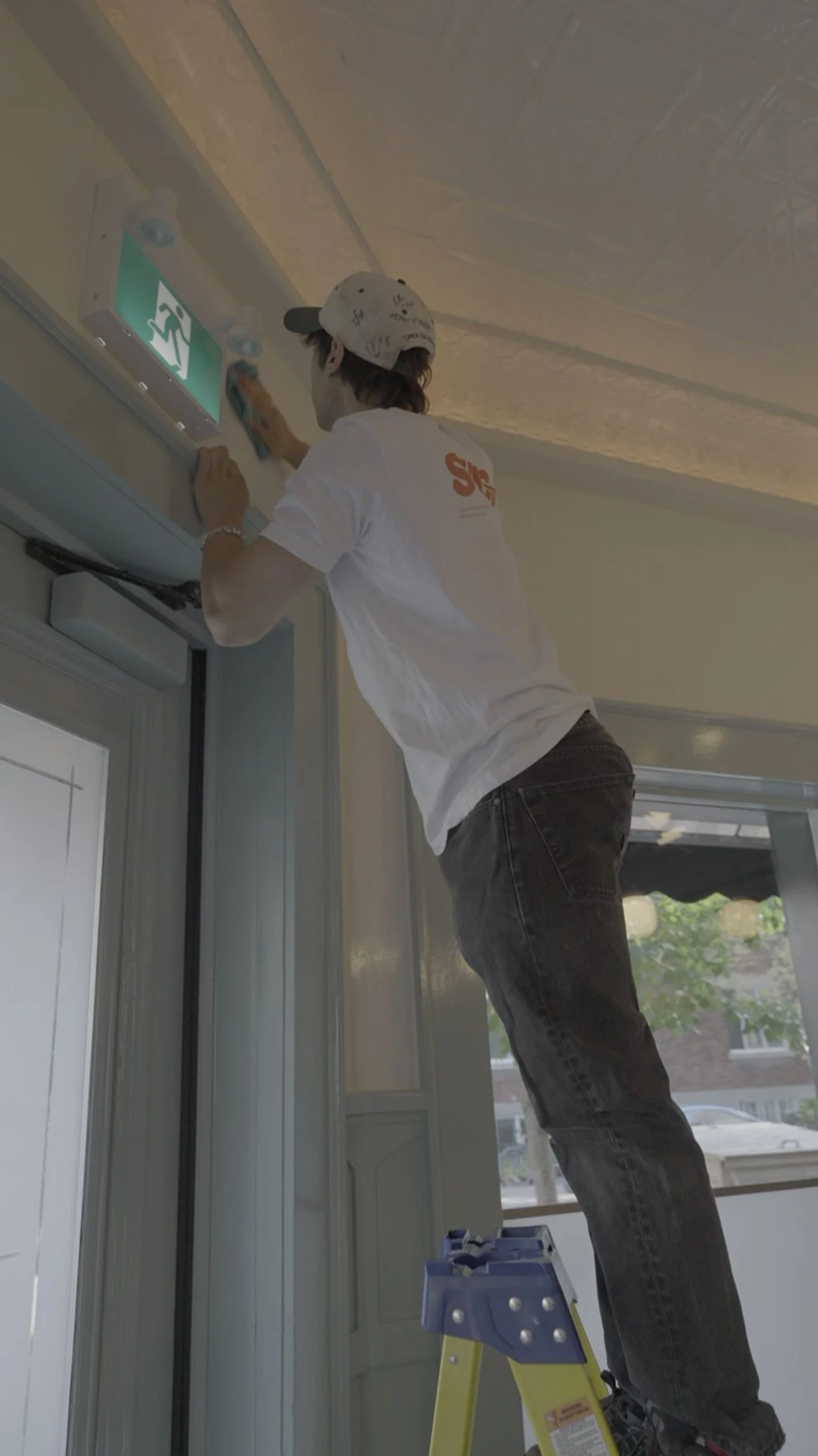 Person standing on a small ladder, cleaning or inspecting an emergency exit sign on the wall near the ceiling.