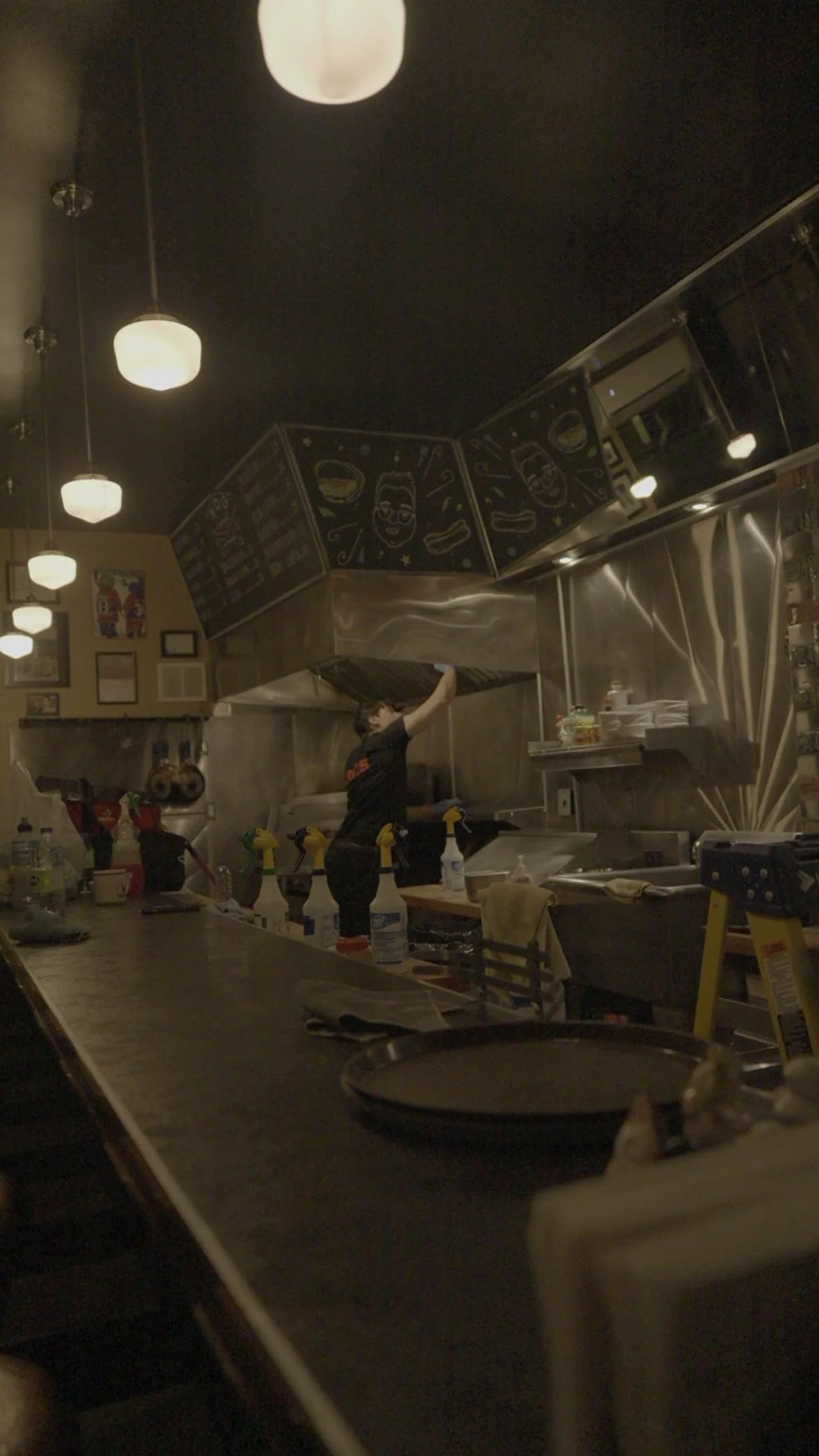 A person cleaning the stainless steel hood in a restaurant kitchen with black walls and hanging lights.