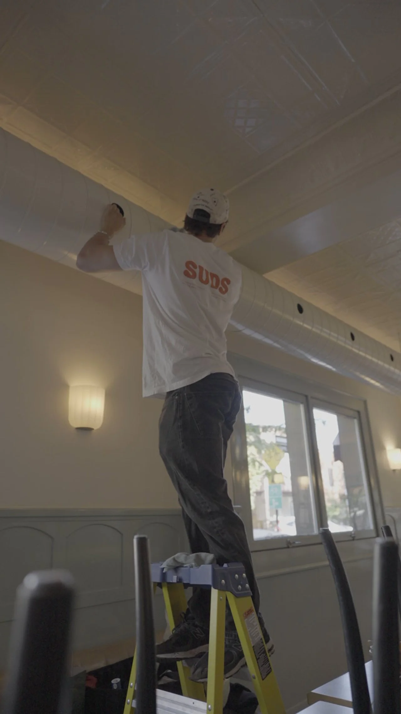 A man standing on a yellow ladder, working on a ceiling in a room with beige walls and large windows, wearing a white t-shirt with 'SUDS' printed on the back and a white baseball cap.