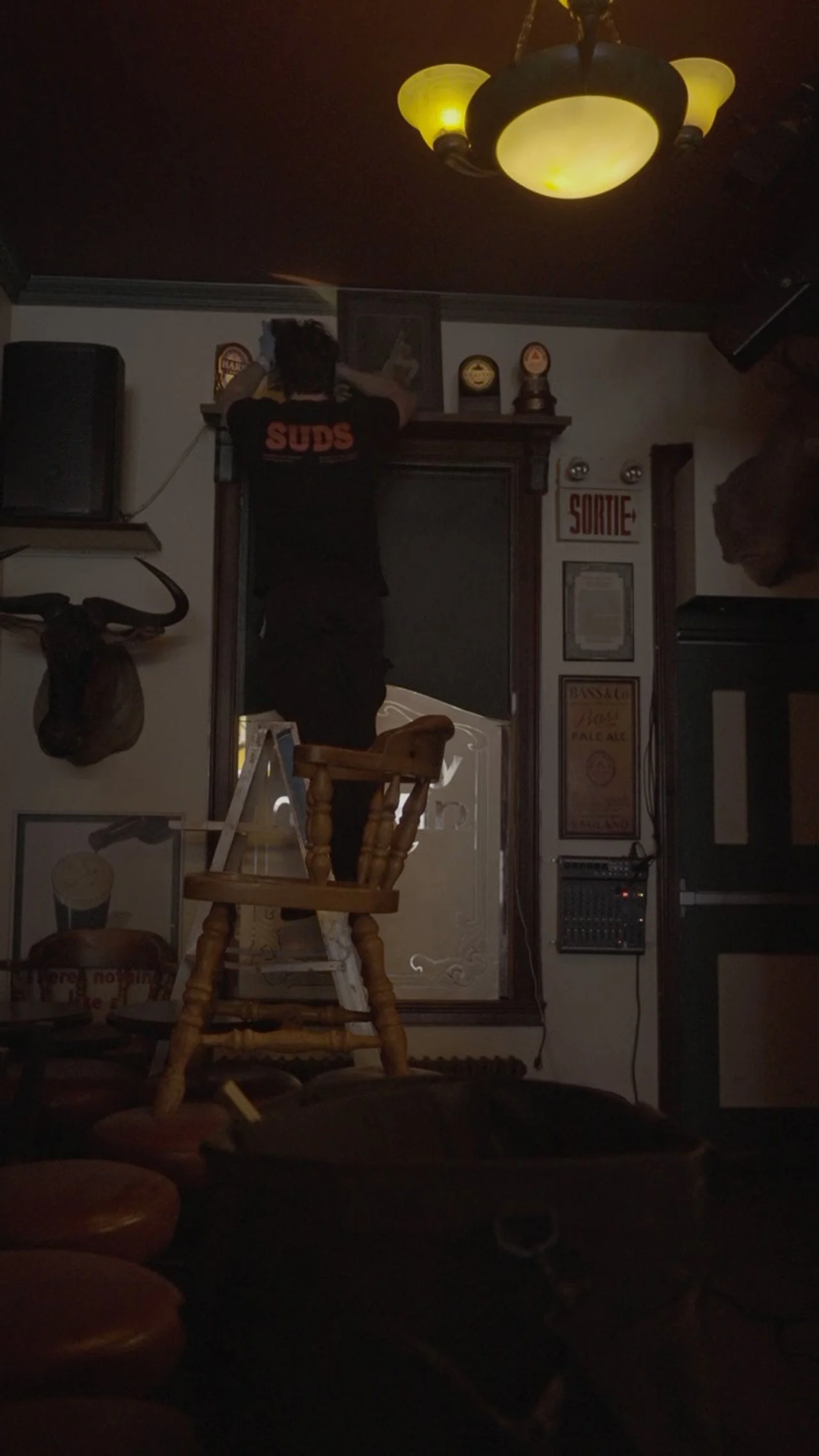 A man standing on a wooden chair, reaching up to a high shelf in a dimly lit room decorated with various signs and trophies.