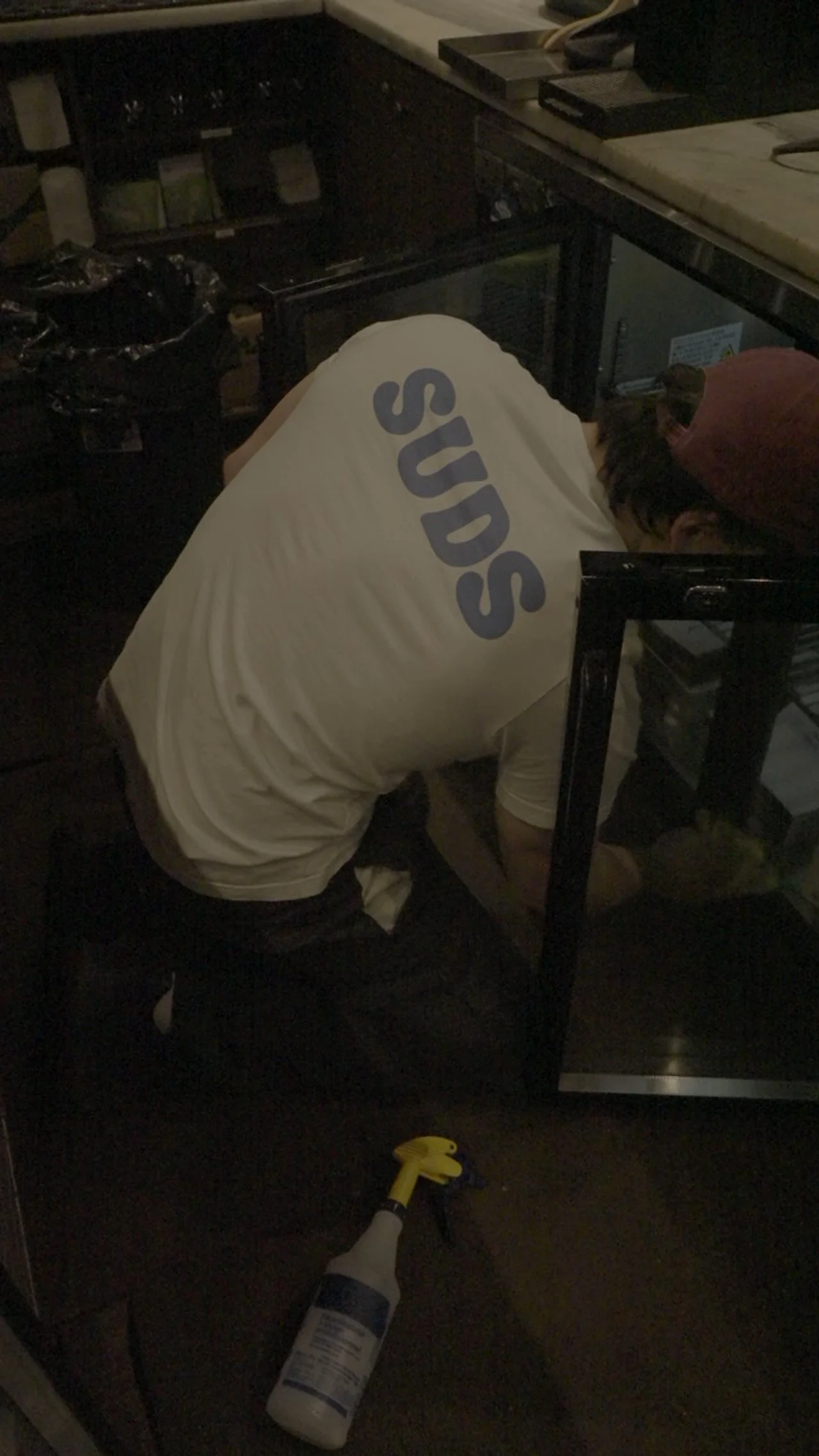 Person wearing a white shirt with the word 'SUDS' on the back, kneeling next to a counter, cleaning or repairing. There is a spray bottle on the floor in front of them.