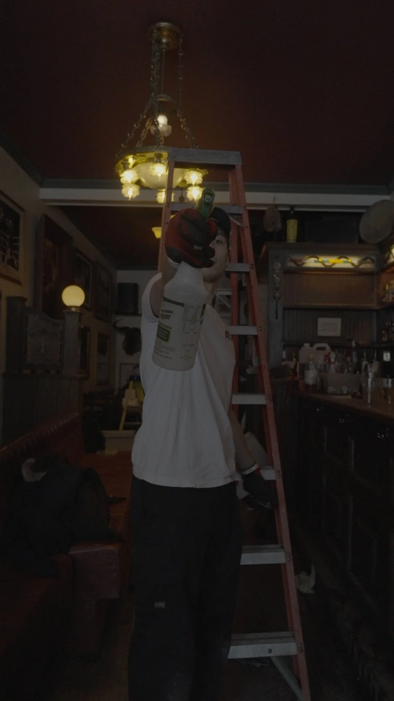 Person standing on a ladder, holding a drill, working on a ceiling light fixture inside a room that appears to be a restaurant or bar.