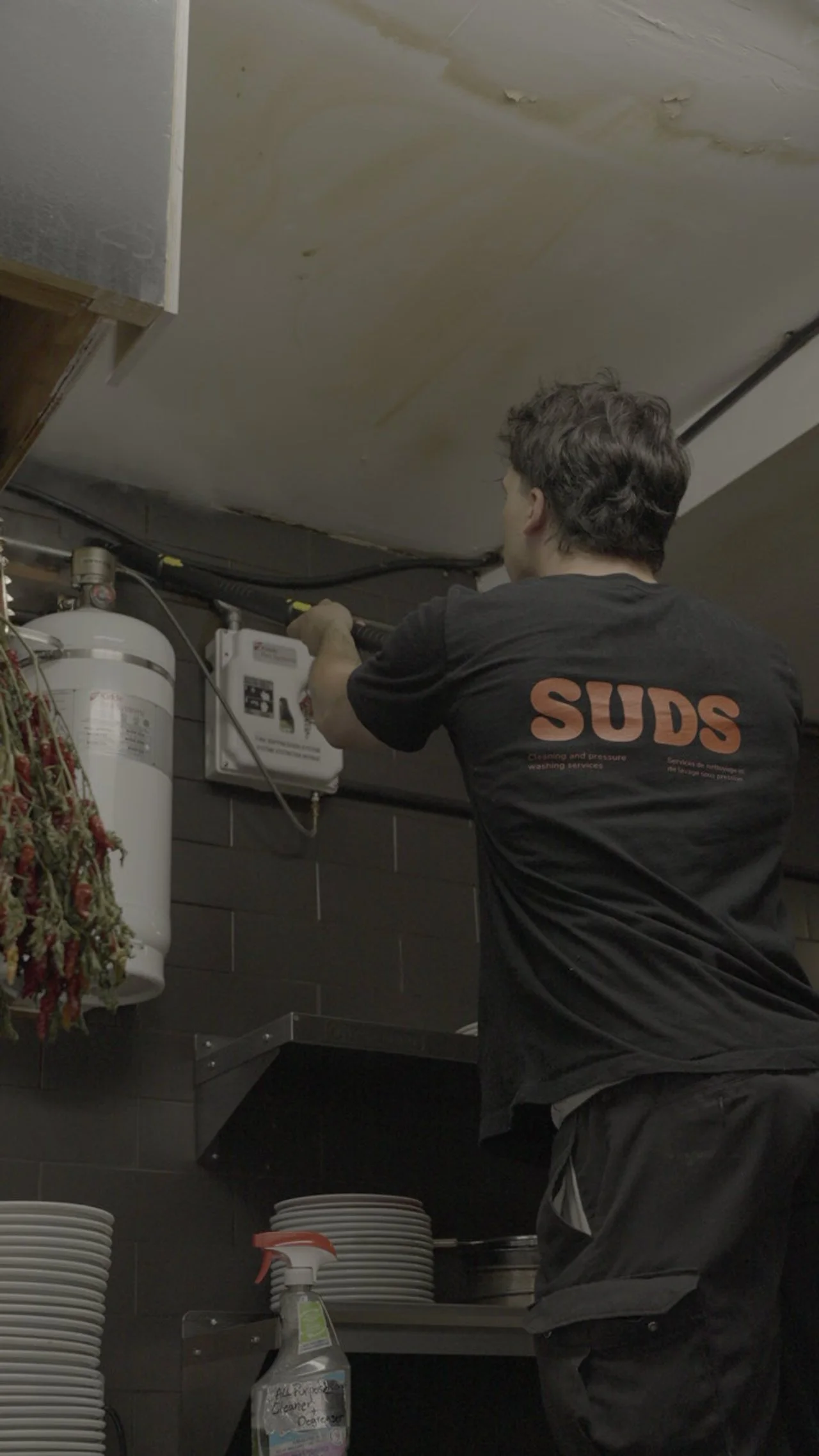 A person wearing a black t-shirt with the word  SUDS in orange letters is working on plumbing or electrical wiring on a ceiling, with a water heater and shelves with plates nearby.