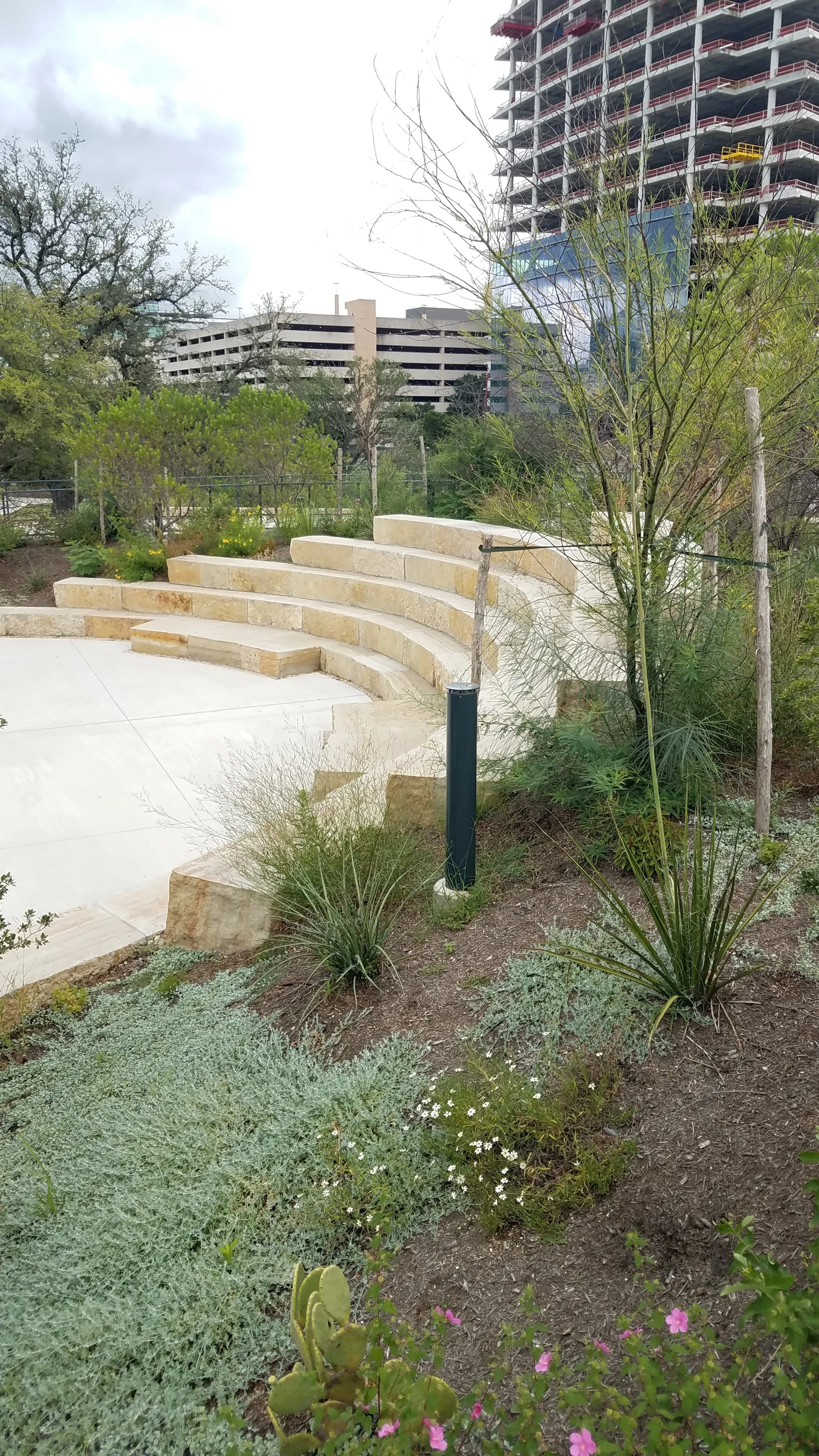 A small outdoor amphitheater with a stone semicircular stage and tiered stone seating, surrounded by desert plants, trees, and a landscaped garden, with a tall construction building and a parking garage in the background.