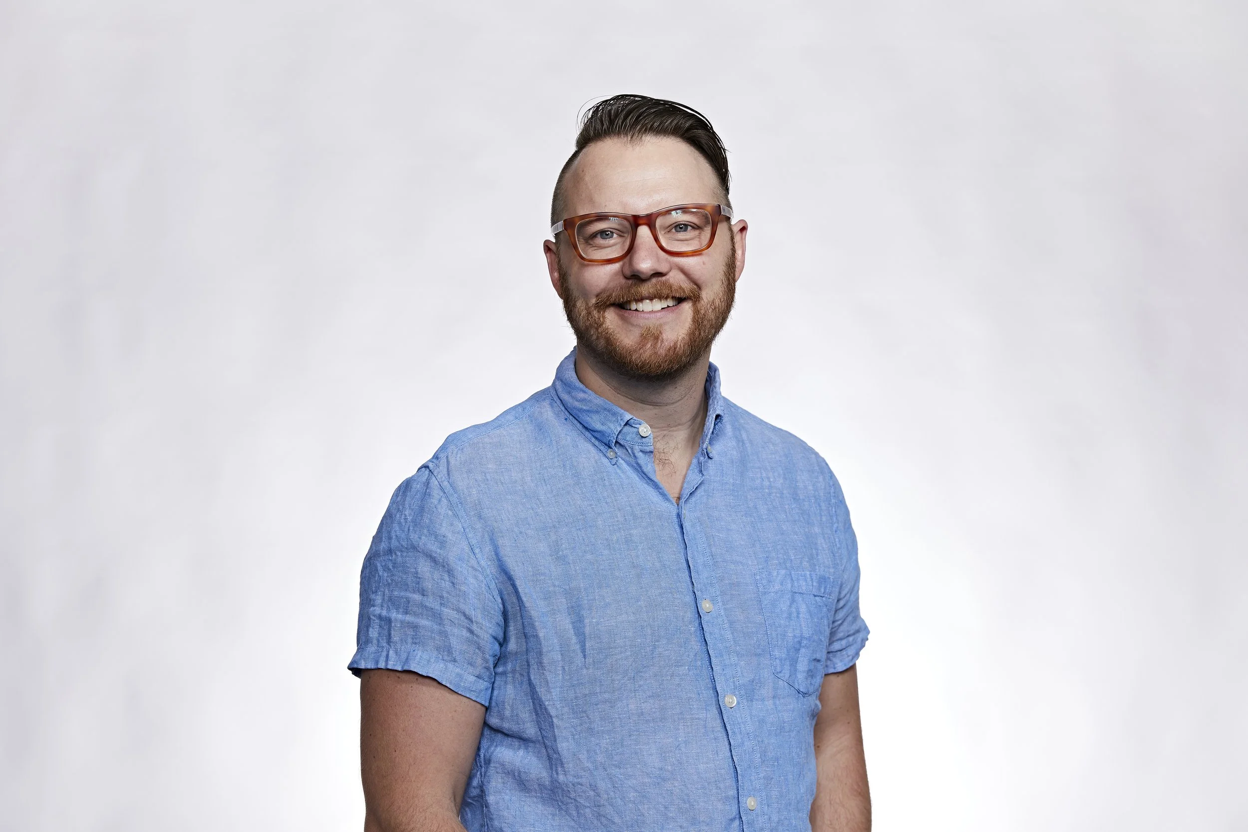 Smiling man with glasses, beard, wearing a blue short-sleeve shirt, standing against a white background.