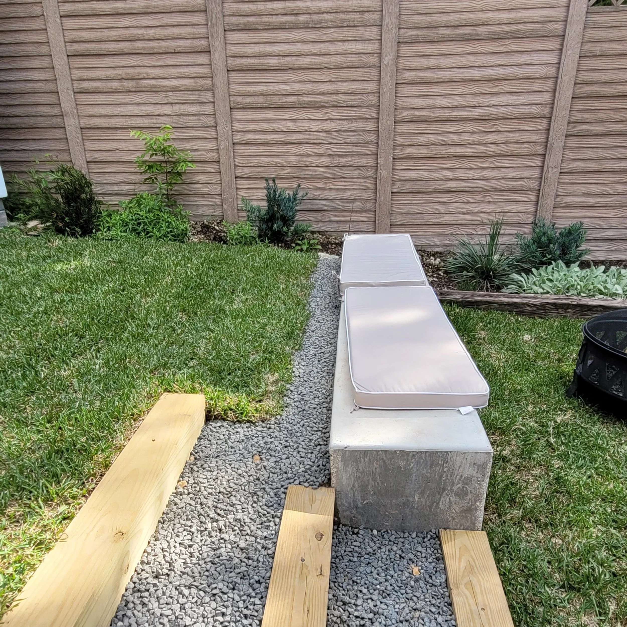 Backyard garden with wooden pathway, concrete block bench with cushions, green grass, and plants near a wooden fence.