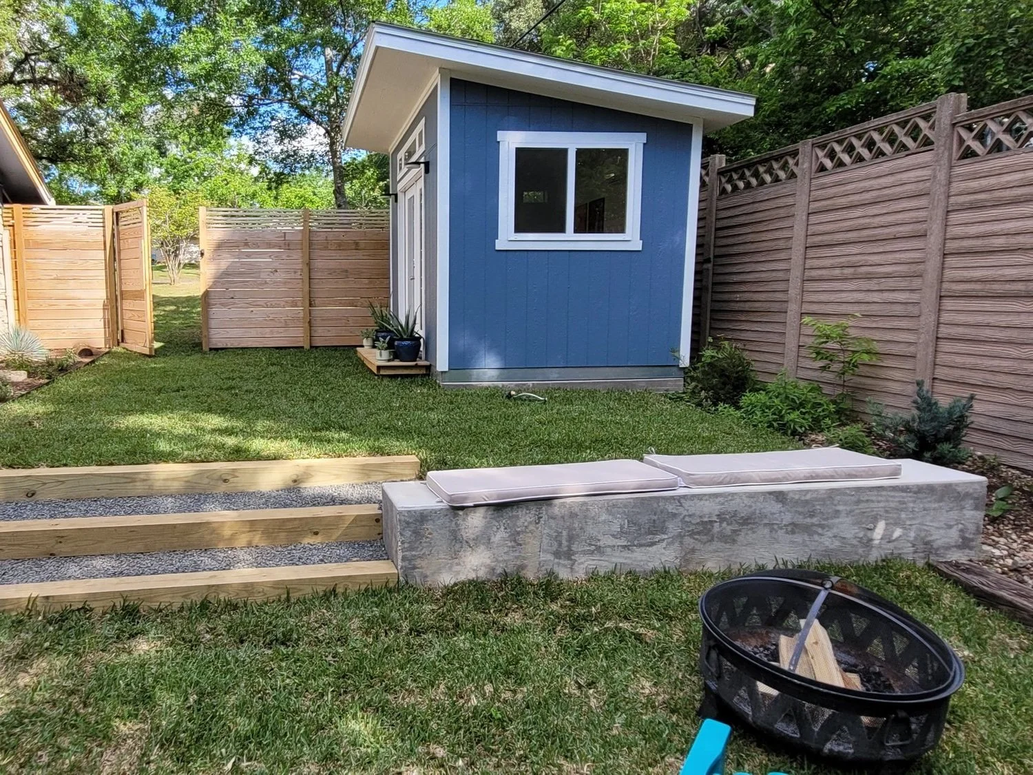 Backyard with a small blue shed, wooden fence, lawn, and garden plants, with seating cushions on a concrete bench and a black portable fire pit in the foreground.