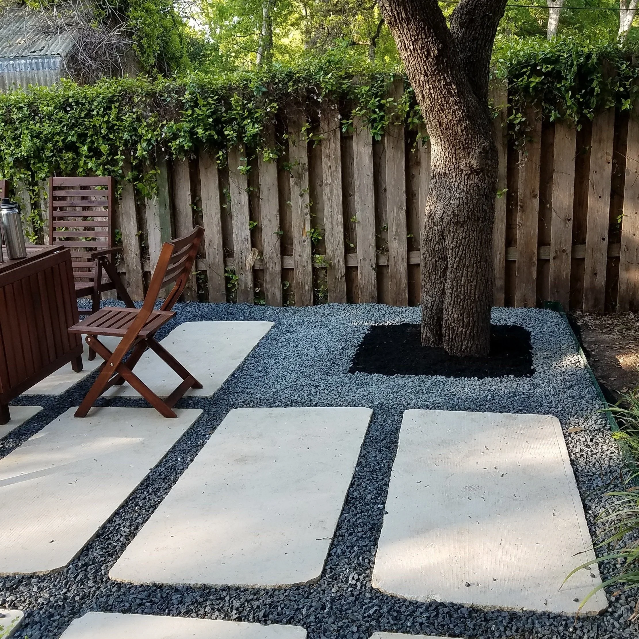 A backyard patio with large concrete stepping stones on a gravel surface, a wooden fence with greenery, a tree with mulch around its base, and outdoor wooden chairs and furniture.