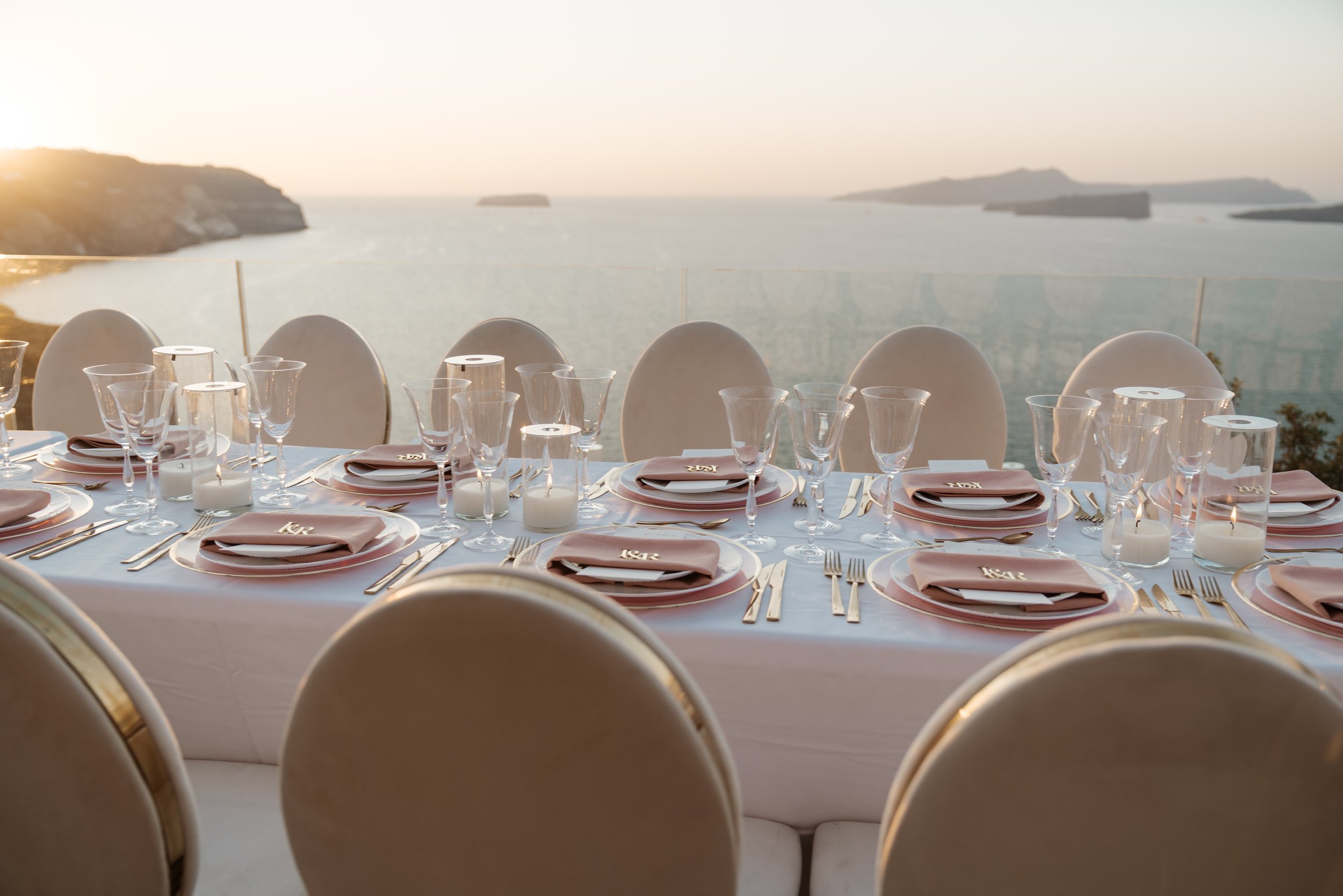 Elegant Santorini wedding dinner setting with blush pink napkins, gold cutlery, candlelight and panoramic Aegean Sea views.