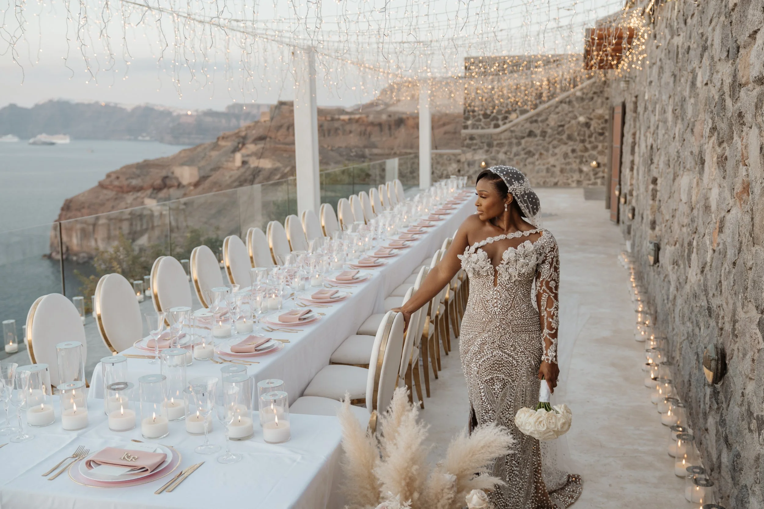 Bride in an intricately embellished gown beside a candlelit wedding reception table overlooking the sea in Santorini, Greece.