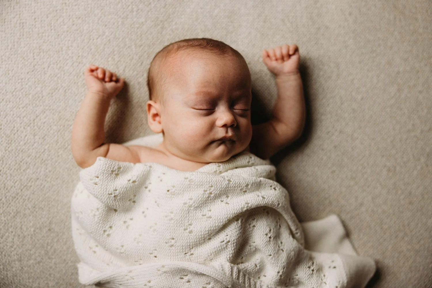 A newborn baby sleeping peacefully on a beige surface, wrapped in a white knitted blanket, with arms raised and eyes closed.