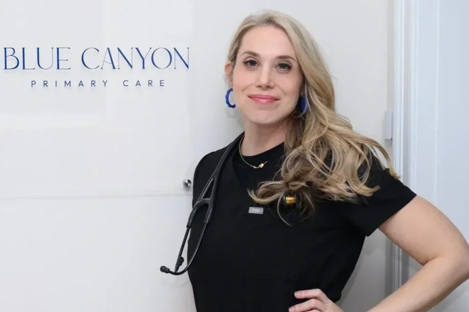 A woman standing in front of a wall with a "Blue Canyon Primary Care" sign, wearing a black medical uniform and a stethoscope around her neck, smiling with one hand on her hip.